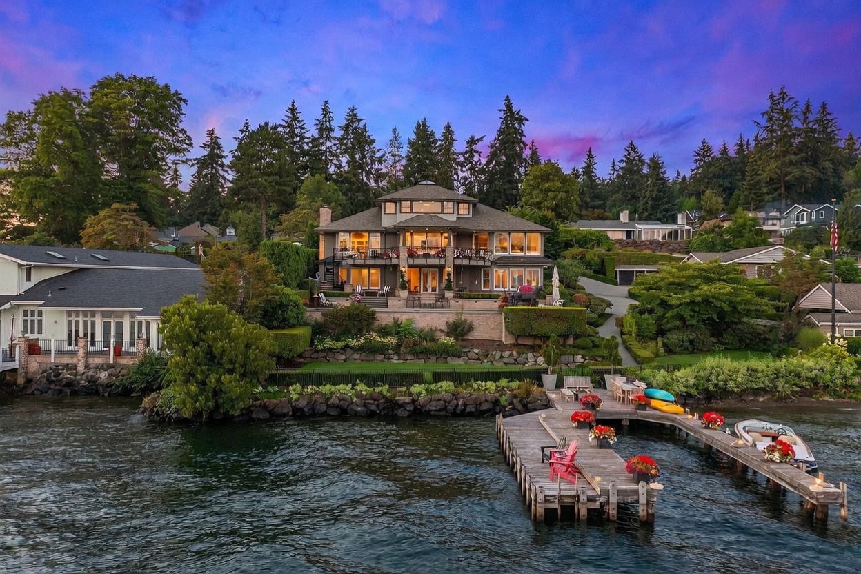 This stunning waterfront property features a multi-level residence with expansive decks and balconies overlooking a private dock on the lake. The home is nestled into a lush, landscaped hillside with stone retaining walls and mature trees, creating a serene and private atmosphere. The twilight sky provides a dramatic backdrop, highlighting the warm interior lighting and the inviting outdoor living spaces.