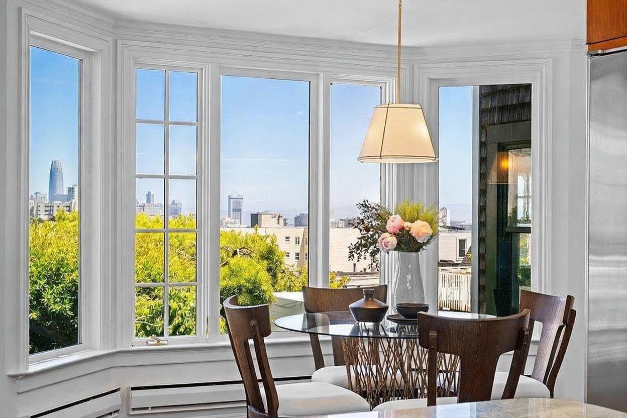 This bright dining area features a glass-topped table surrounded by four wooden chairs with light-colored upholstery. A large, multi-pane bay window provides an expansive view of the city skyline, filling the space with natural light. A simple pendant light hangs above the table, which is decorated with a vase of flowers and a decorative bowl, creating an inviting and airy atmosphere.