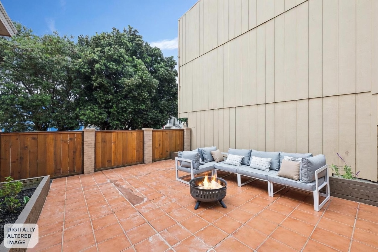 This outdoor patio space features terracotta-colored tiles and a modern sectional sofa arranged around a circular fire pit. The area is enclosed by a wooden fence with brick pillars, providing a private setting against the side of a building. The perspective is a wide-angle shot from the patio level, capturing the inviting lounge area and the surrounding outdoor environment.