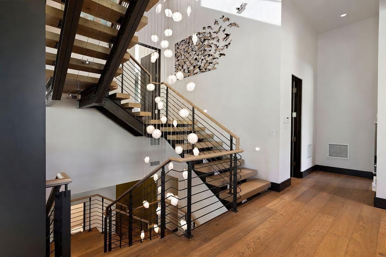 This striking interior space features a grand, multi-level staircase with light wood treads and a modern black metal railing. A dramatic, cascading chandelier with numerous glowing orb lights serves as the focal point, hanging vertically through the open stairwell. The walls are painted a crisp white, complemented by a large, textured wall art installation and warm-toned wood flooring that extends into the adjacent hallway.