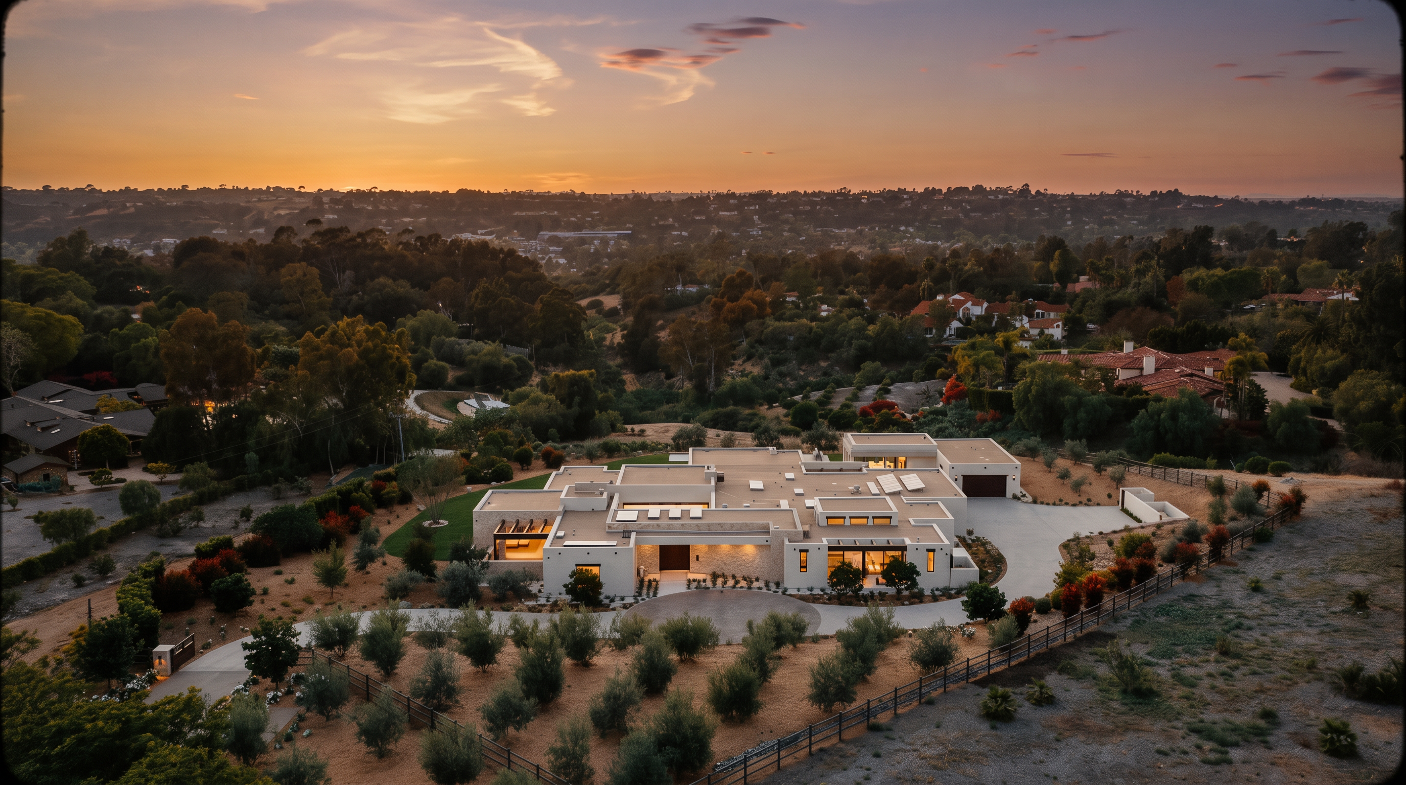 This stunning aerial perspective captures a sprawling, modern single-story estate set against a vibrant sunset sky. The residence features a low-profile, contemporary architectural design with a flat roofline, expansive glass walls, and a neutral-toned exterior that blends seamlessly into the surrounding landscape. A circular driveway leads to the main entrance, while the property is framed by manicured grounds, olive trees, and a vast, rolling hillside vista.