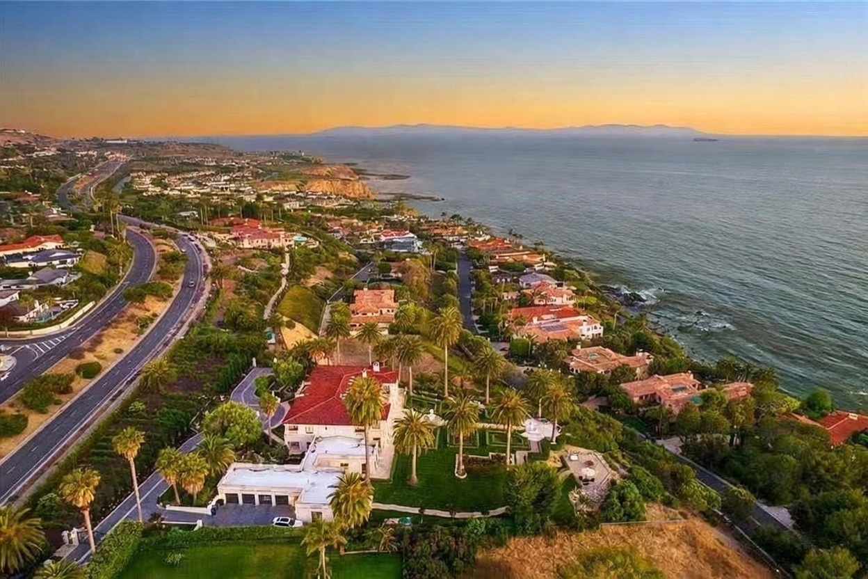 This high-angle aerial perspective captures a sprawling coastal estate nestled along a scenic cliffside overlooking the ocean. The property features a large residence with a red-tiled roof, manicured formal gardens, and a private driveway, all set against a backdrop of a winding coastal road and distant mountains at sunset. The expansive view highlights the prime location, emphasizing the seamless integration of luxury living with the natural beauty of the shoreline.