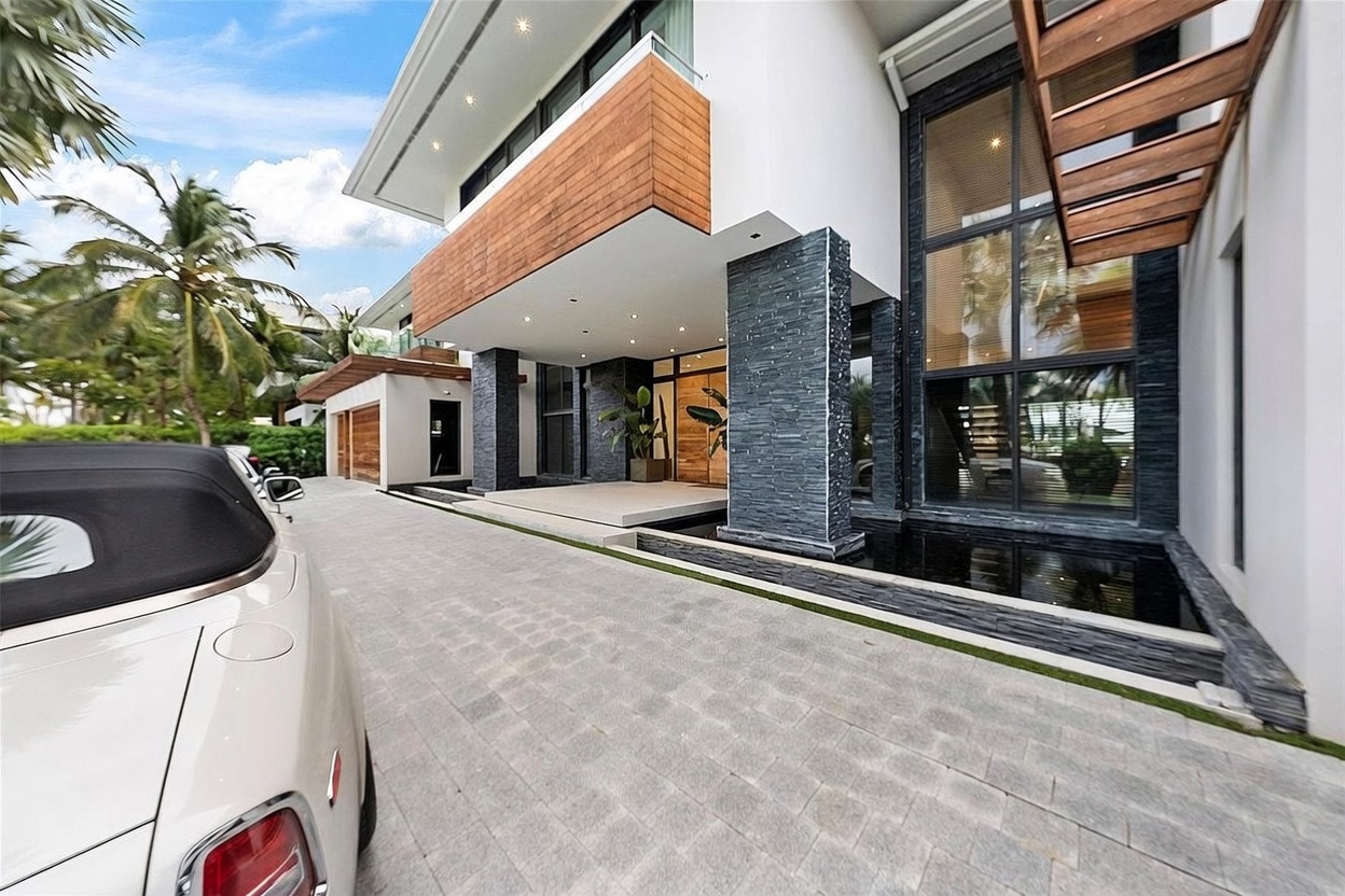 This modern luxury residence features a striking entryway characterized by a cantilevered upper level clad in warm-toned wood siding. The ground level showcases a dramatic combination of dark stone pillars, a reflective water feature, and large glass panels that offer a glimpse into the interior. A paved driveway leads up to the entrance, with a portion of a luxury vehicle visible in the foreground, emphasizing the high-end nature of the property.