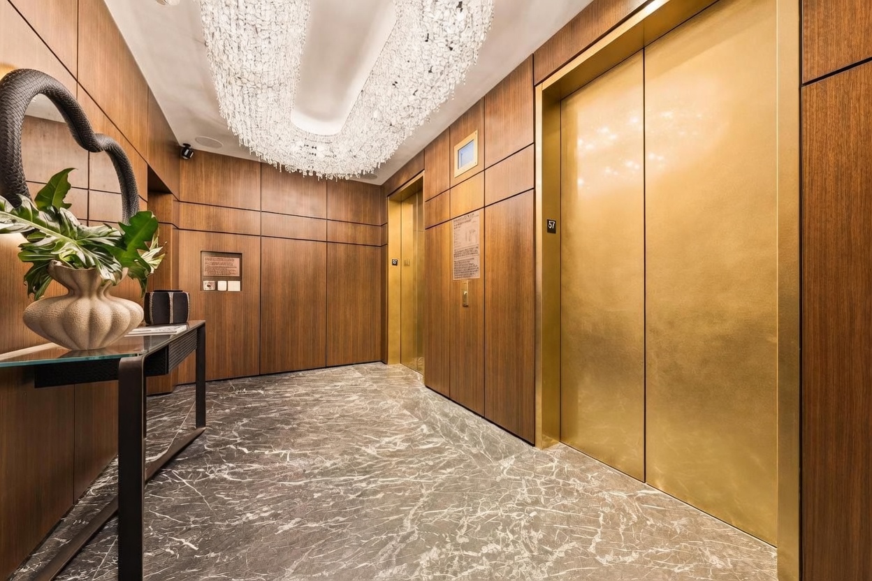 This elegant elevator lobby features rich wood-paneled walls and a polished stone floor with intricate veining. A dramatic, multi-tiered crystal chandelier hangs from the ceiling, casting a warm glow over the space, while a sleek console table with a decorative plant adds a touch of sophistication. The scene is anchored by two prominent gold-toned elevator doors, creating a luxurious and high-end atmosphere.