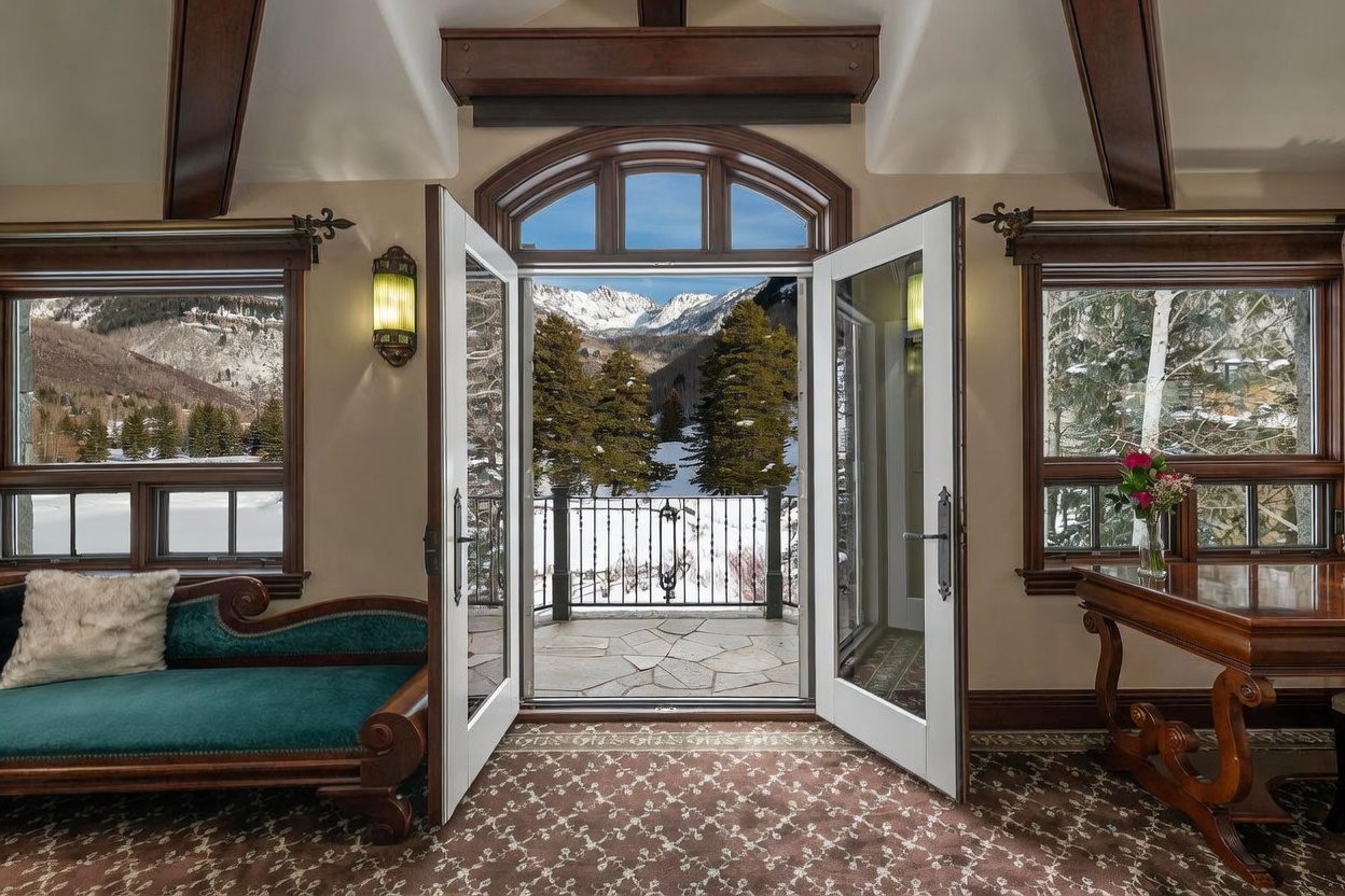 This elegant interior space features a plush teal chaise lounge and a dark wood desk, set against a backdrop of rich, patterned carpeting and exposed wooden ceiling beams. The room is centered around a pair of open French doors that frame a breathtaking, unobstructed view of snow-capped mountains and evergreen trees on a private balcony. The combination of warm, traditional furnishings and the dramatic, expansive mountain vista creates a sophisticated and serene atmosphere.