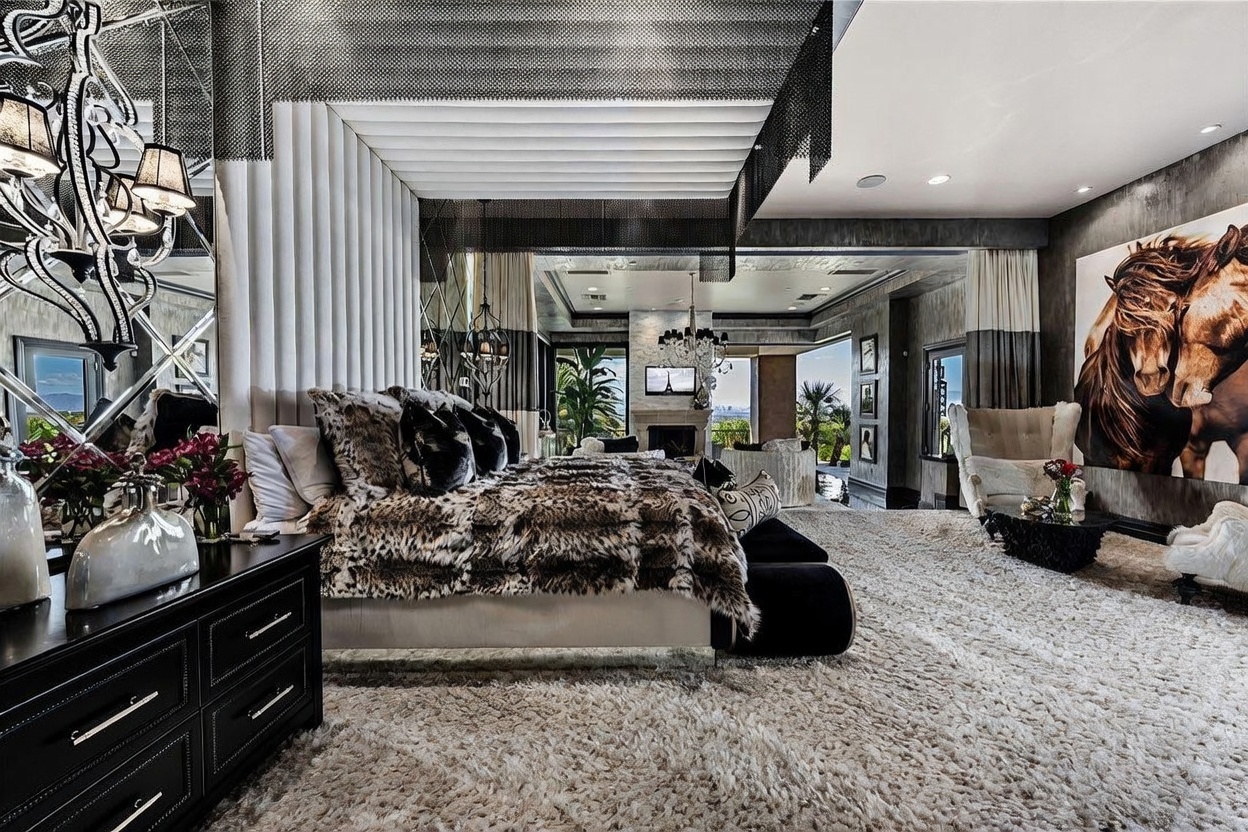 This opulent primary bedroom features a dramatic canopy bed draped in animal-print textiles, set against a backdrop of mirrored walls and a textured ceiling treatment. The room flows into an expansive seating area with a fireplace and large windows that frame a scenic outdoor view. A large, striking horse portrait serves as a focal point on the wall, contributing to the room's luxurious and bold aesthetic.