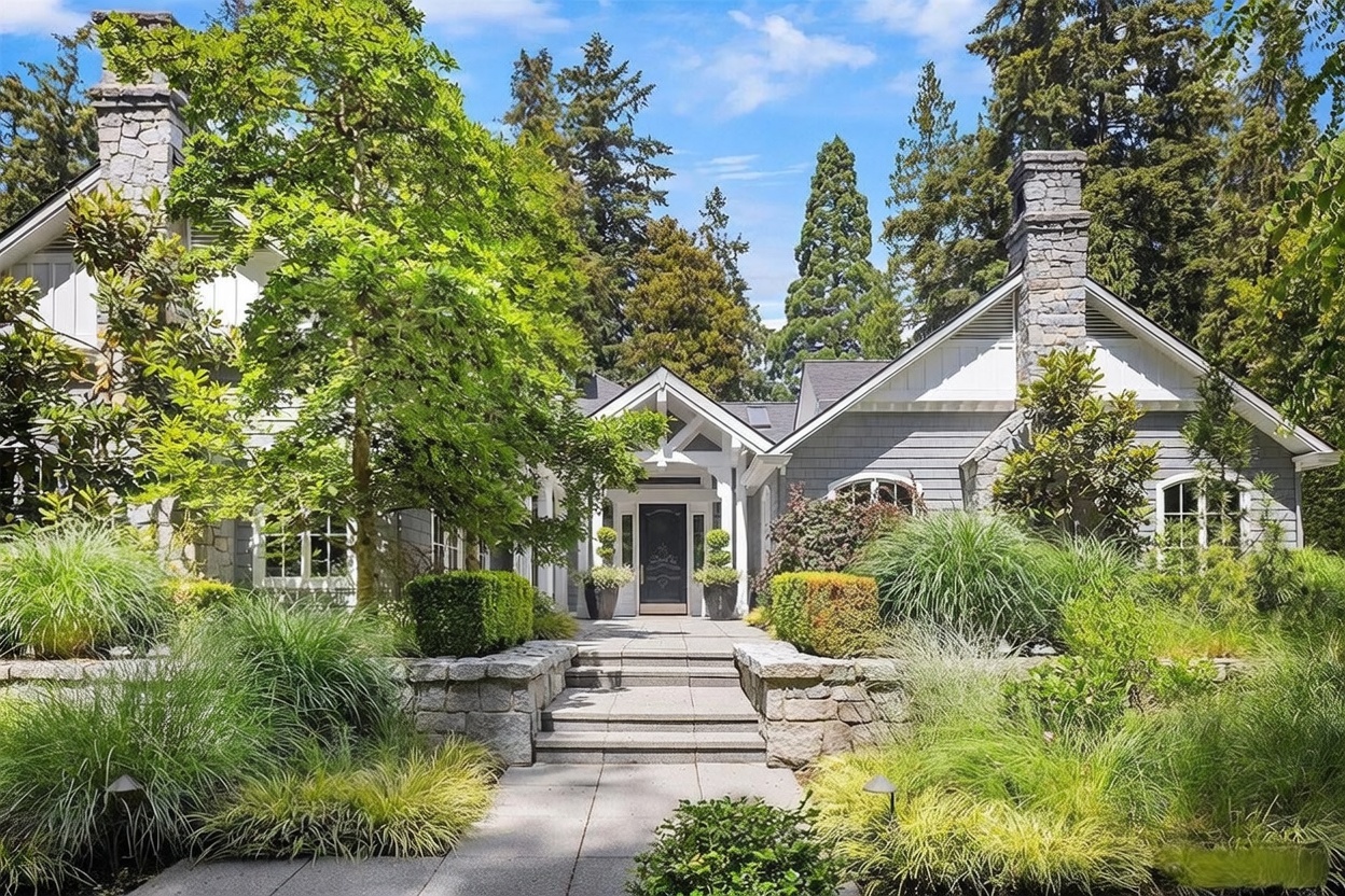 This elegant residence features a classic architectural style with gray shingle siding, white trim, and prominent stone-clad chimney breasts on either side. A stone-paved walkway leads up a set of steps to a welcoming front entrance framed by a covered porch and lush, manicured landscaping. The scene is set against a backdrop of mature evergreen trees under a bright, clear blue sky, creating a serene and inviting curb appeal.