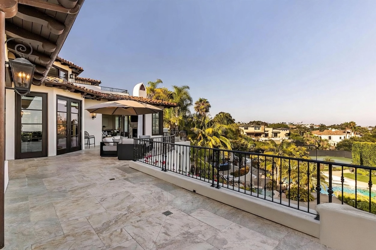 This expansive, elevated stone-tiled balcony offers a luxurious outdoor living space with a wrought-iron railing overlooking a lush landscape and a swimming pool below. The area is furnished with comfortable lounge seating under a large beige umbrella, creating a perfect spot for relaxation while enjoying the panoramic views of the surrounding neighborhood. The architecture features classic Mediterranean-inspired elements, including a tiled roof and a prominent wall-mounted lantern, providing a sophisticated and inviting atmosphere.