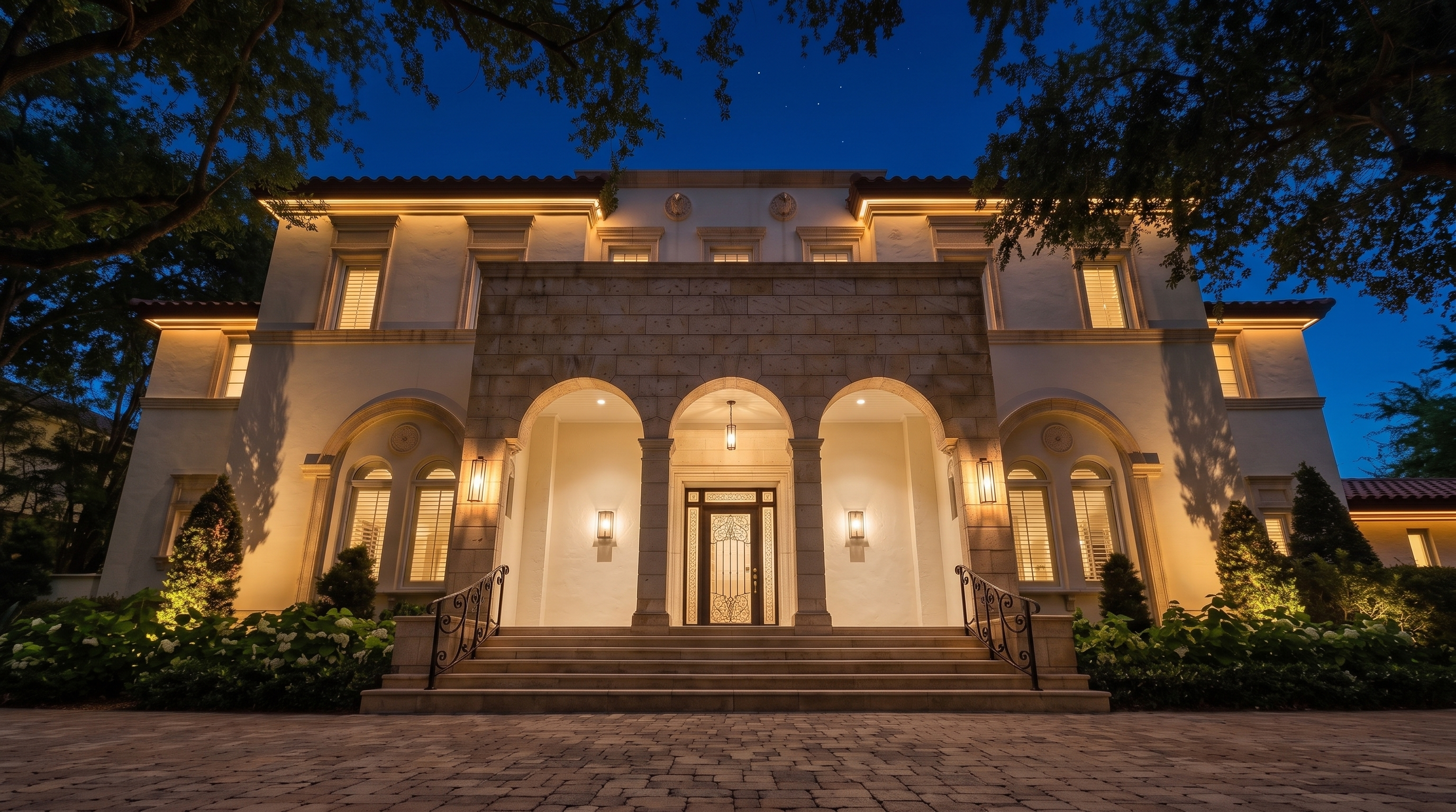 This grand, Mediterranean-inspired estate features a symmetrical facade with a prominent, stone-clad triple-arch entryway and elegant exterior lighting at dusk. The property showcases meticulous landscaping, a paved motor court, and a warm, inviting porch that leads to a detailed custom front door. Its sophisticated architectural design and warm, ambient illumination create a luxurious and impressive first impression.