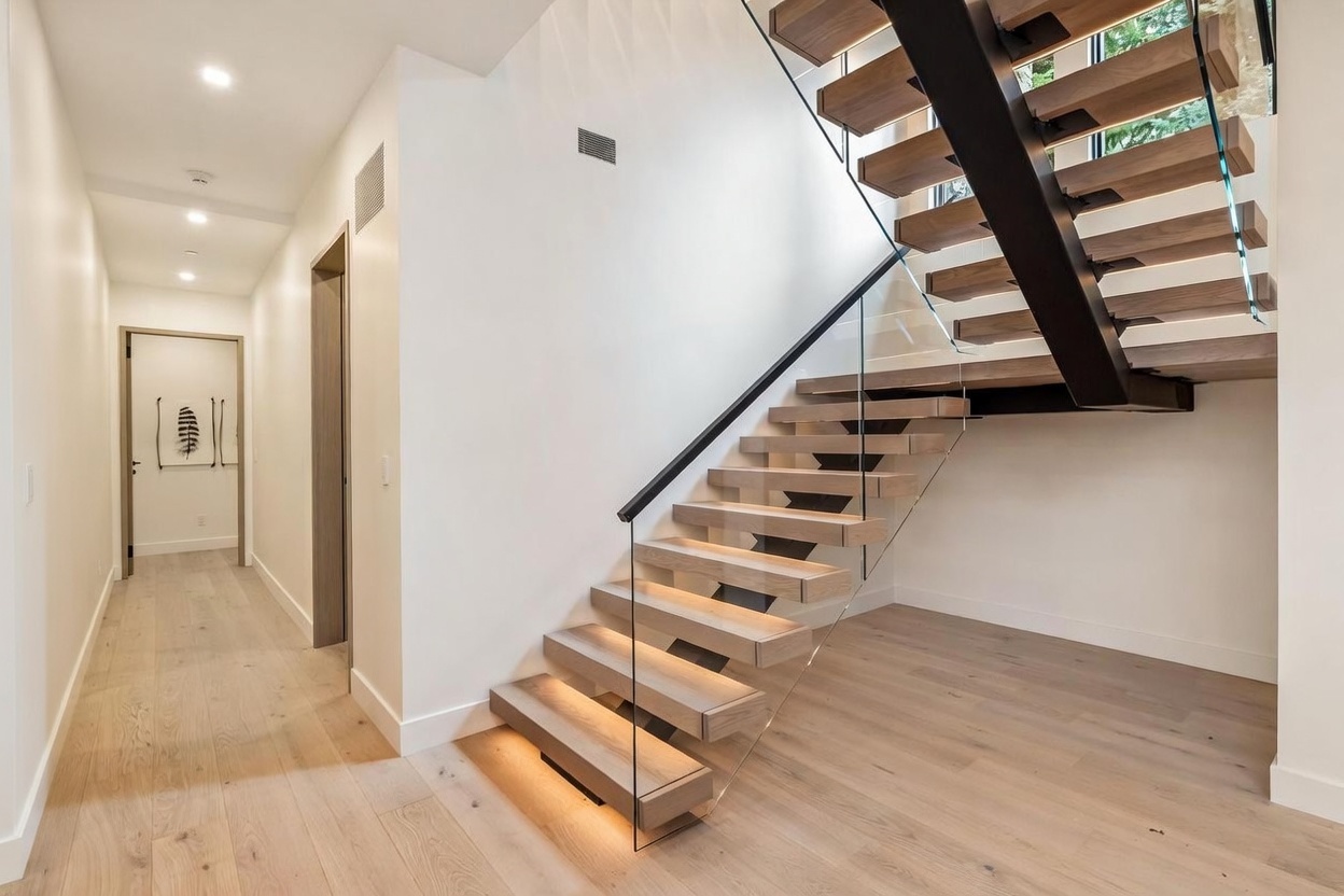 This modern interior space features a striking floating staircase with light wood treads and a sleek glass railing. The hallway leads to a doorway, with light wood flooring extending throughout the area and integrated lighting beneath the stair treads. The clean, minimalist aesthetic is enhanced by bright white walls and a neutral color palette.
