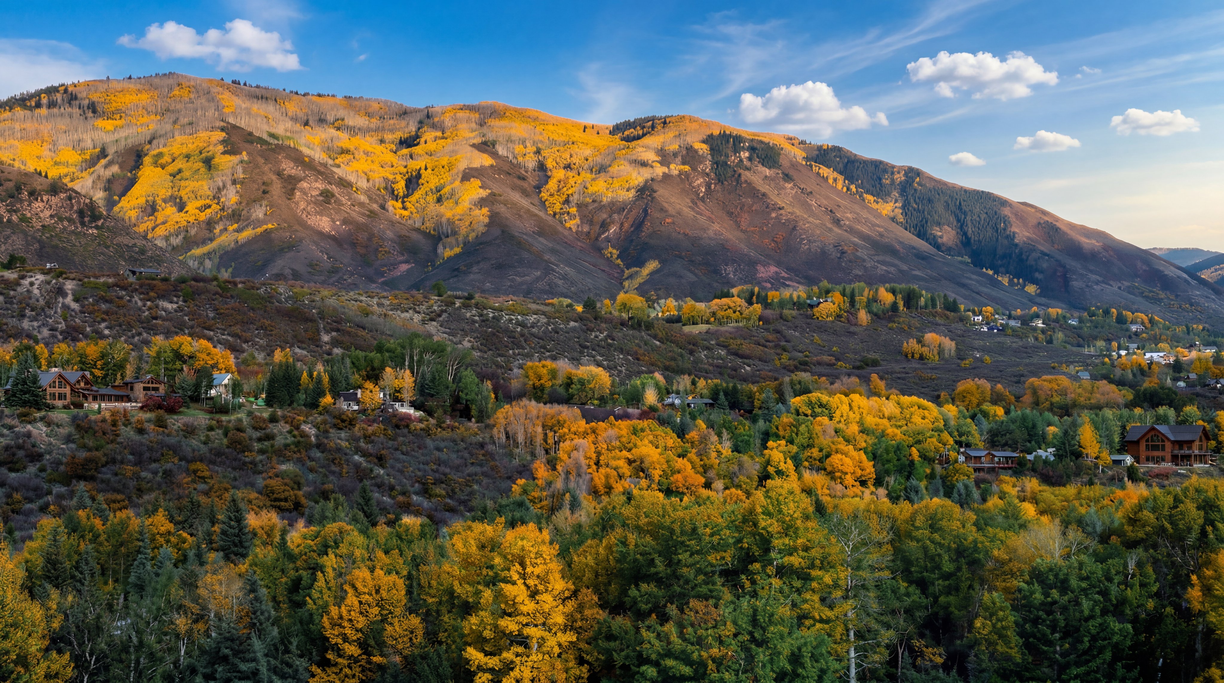 This wide-angle aerial view captures a sprawling mountainous landscape during the peak of autumn, characterized by vibrant yellow aspen trees contrasting against dark, rugged slopes. The scene showcases the natural beauty of the region, with scattered residential properties nestled among the dense, colorful foliage. The perspective provides a grand, cinematic sense of scale, emphasizing the serene and secluded nature of the location.