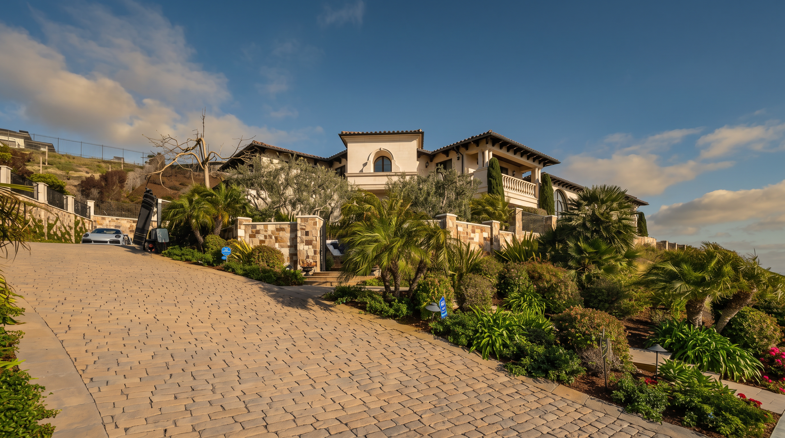This image captures a grand, Mediterranean-style estate from a low-angle perspective, emphasizing its elevated position on a hillside. A long, winding driveway paved with textured stone leads up to the residence, which features a multi-level facade with stucco walls, arched architectural details, and a tiled roof. The property is surrounded by lush, tiered landscaping, including mature palm trees and various shrubs, creating a sense of privacy and luxury.