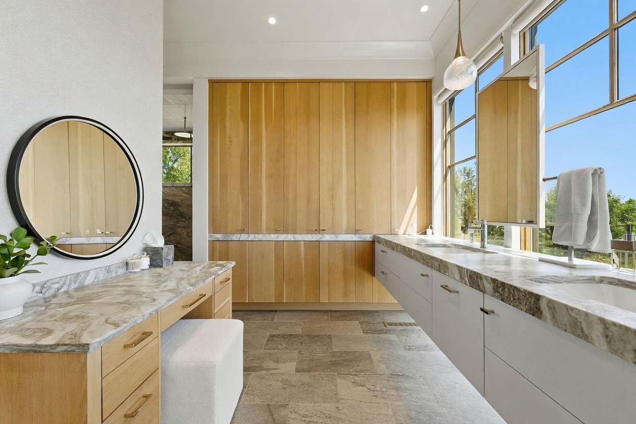 This modern primary bathroom features a dual-vanity setup with light wood cabinetry and a stone countertop. A circular mirror hangs above a dedicated vanity station, while a large rectangular mirror is positioned over the sink area, complemented by a hanging pendant light. The space is brightened by large windows that provide natural light and a view of the outdoors, with stone-look tile flooring completing the clean, contemporary aesthetic.