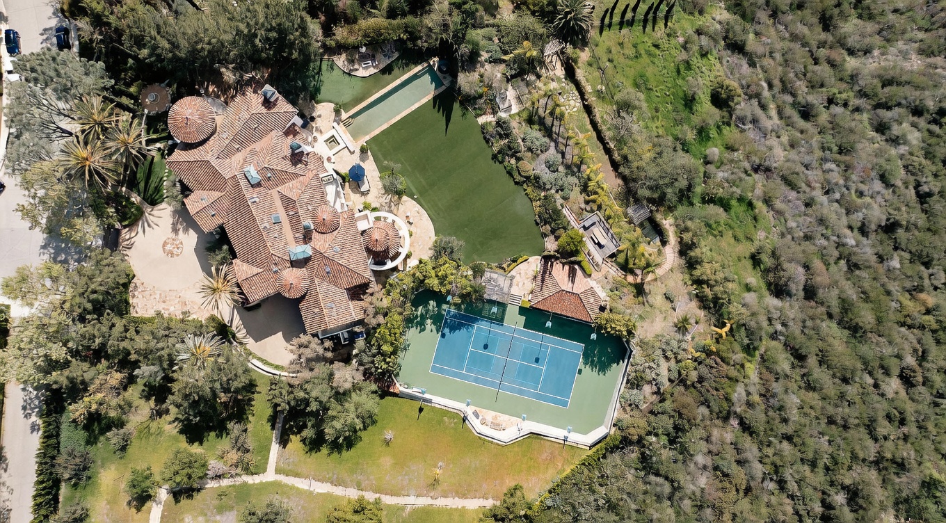 This high-angle aerial view captures a sprawling luxury estate nestled against a hillside, featuring a large Mediterranean-style residence with a complex tiled roof. The property includes a private tennis court, a long rectangular swimming pool, and expansive manicured lawn areas surrounded by mature trees and natural landscape. The perspective provides a comprehensive look at the layout of the grounds, highlighting the seamless integration of recreational amenities and residential architecture.