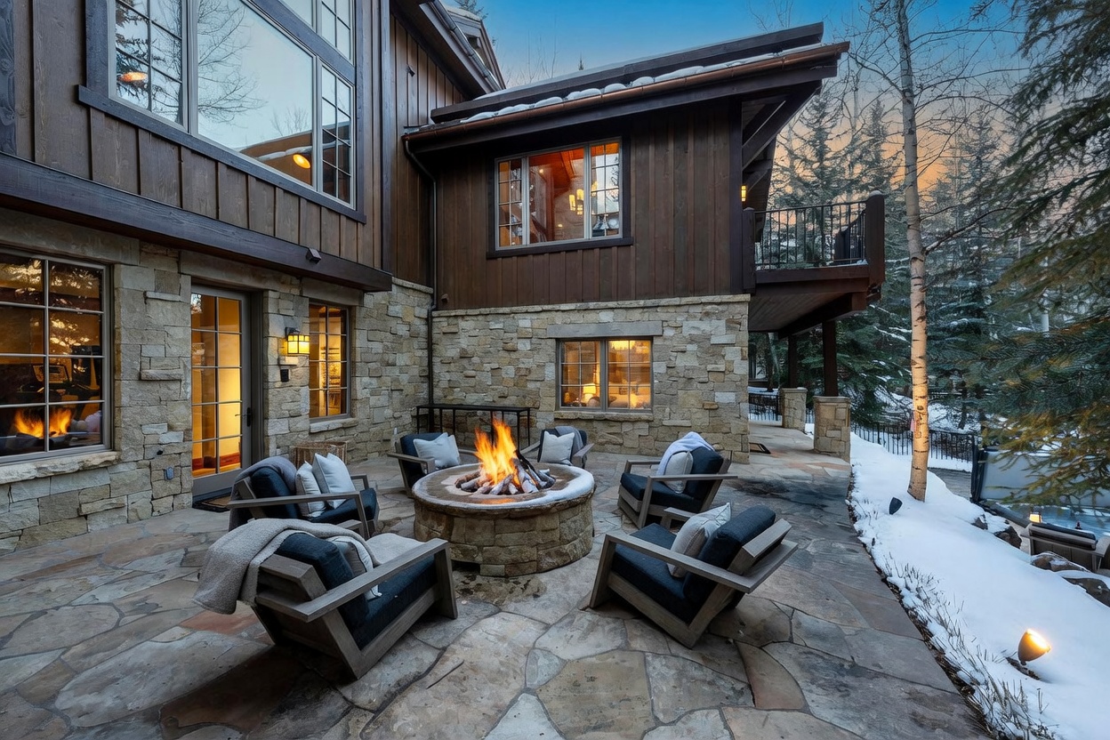 This inviting stone patio features a central circular fire pit surrounded by comfortable outdoor lounge chairs, creating a cozy gathering space. The area is set against a rustic home exterior with wood siding and stone masonry, with snow-dusted trees visible in the background. The scene captures a serene, upscale mountain retreat atmosphere during the twilight hours.