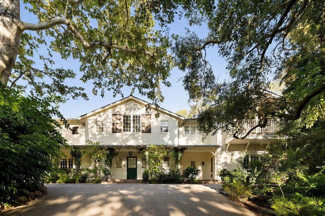 This image captures the front facade of a charming, two-story traditional home nestled under a canopy of mature, leafy trees. The exterior features white vertical siding, a dark green front door, and a covered porch area, creating a welcoming and private atmosphere. The perspective is a straight-on shot from the driveway, emphasizing the home's symmetrical architecture and lush, shaded surroundings.