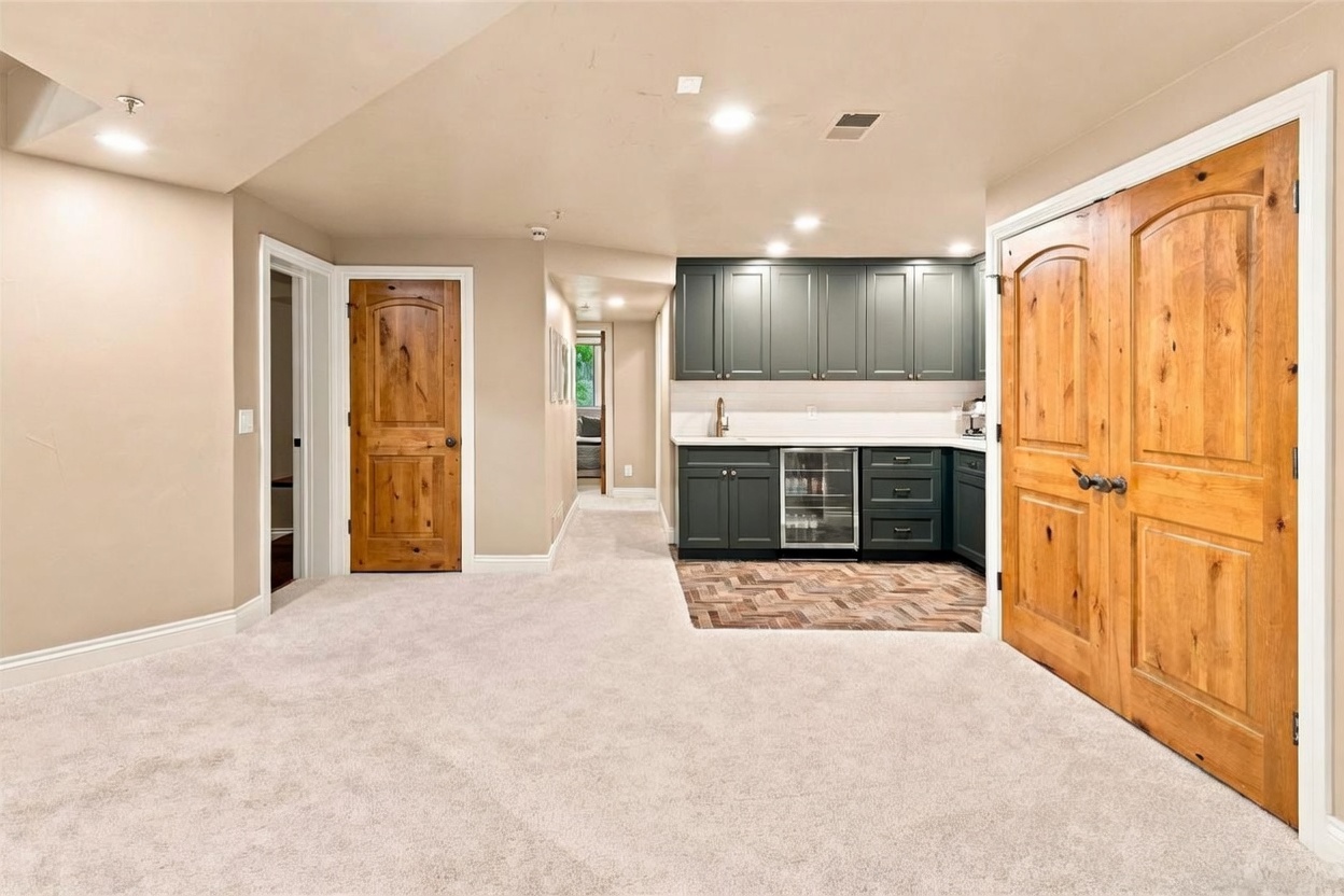 This finished basement area features a wet bar with dark cabinetry, a beverage refrigerator, and a herringbone-patterned tile floor. The space is carpeted and includes double wooden doors leading to an adjacent room, along with a hallway that provides access to further living areas. The neutral wall color and recessed lighting create a clean, versatile space suitable for entertaining.