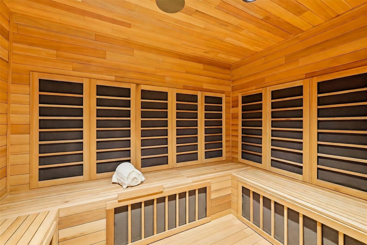 This image showcases a custom-built home sauna constructed entirely from light-toned wood paneling. The space features tiered bench seating and integrated heating panels behind protective grilles, creating a warm and inviting atmosphere. A single white towel rests on the bench, emphasizing the room's purpose as a dedicated wellness retreat.