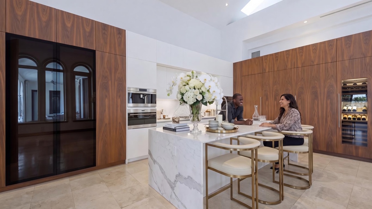 This sophisticated, modern kitchen showcases a blend of sleek white cabinets and rich wood paneling, creating a warm yet contemporary aesthetic. A large marble-topped island serves as the centerpiece, complete with elegant bar seating, while high-end built-in appliances and a dedicated wine refrigeration unit add to its functional luxury. The space is bright and airy, enhanced by minimalist lighting and large, clean surfaces that project a high-end, professionally designed atmosphere.