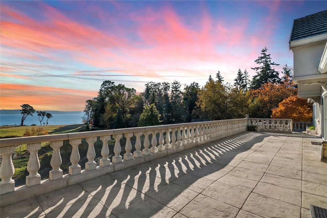 This expansive stone-paved balcony offers a commanding view of a serene body of water and a lush, tree-lined landscape under a vibrant sunset sky. The space is defined by a classic stone balustrade that runs the length of the terrace, casting long, dramatic shadows across the floor. The perspective captures the transition from the structured, elegant architecture of the home to the natural beauty of the horizon.