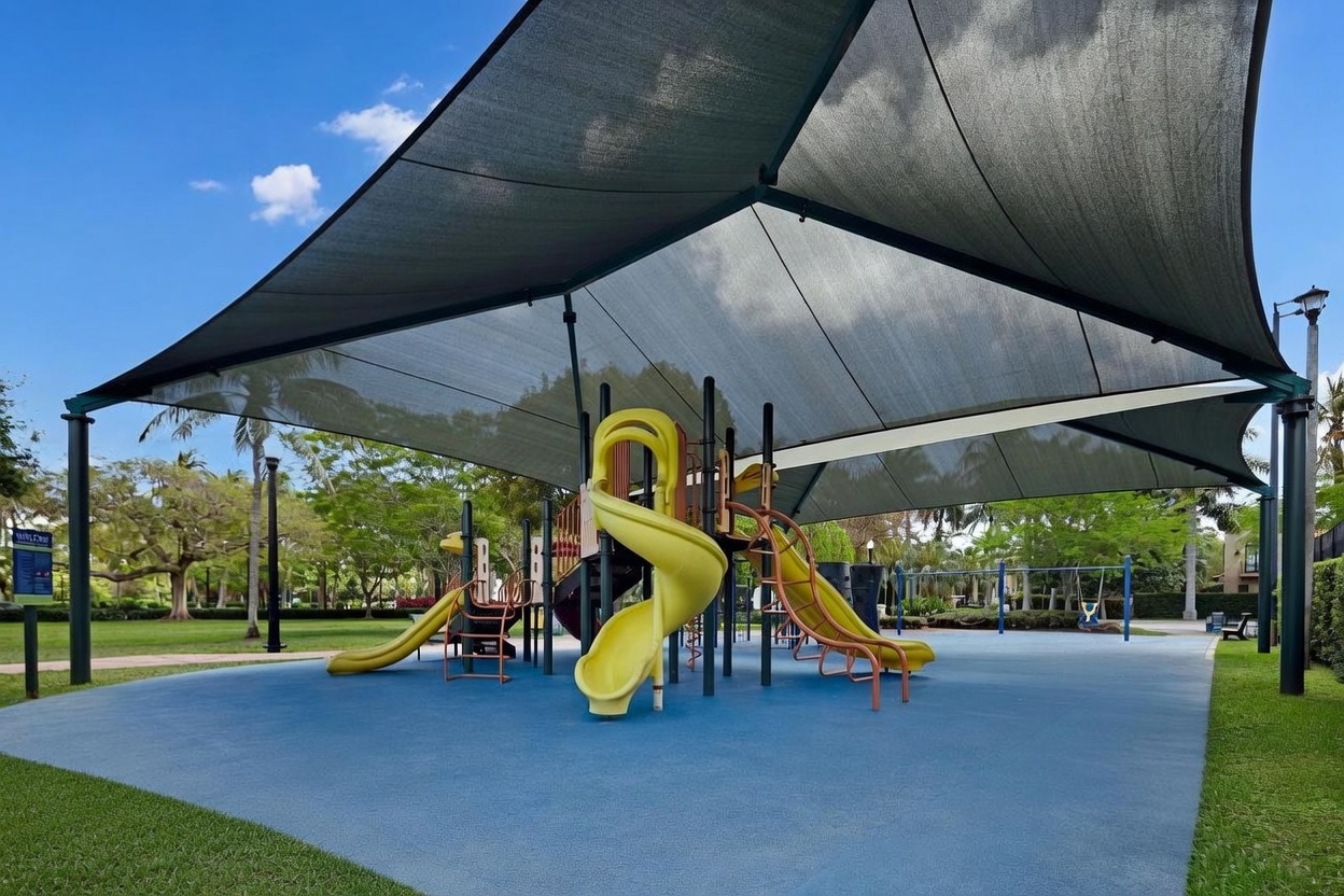 This image showcases a vibrant community playground featuring a multi-level play structure with yellow slides, all protected by a large, modern shade canopy. The play area is set on a blue, impact-absorbing surface and is surrounded by lush green trees and manicured landscaping. The perspective is a wide, eye-level shot that captures the inviting and well-maintained recreational space.