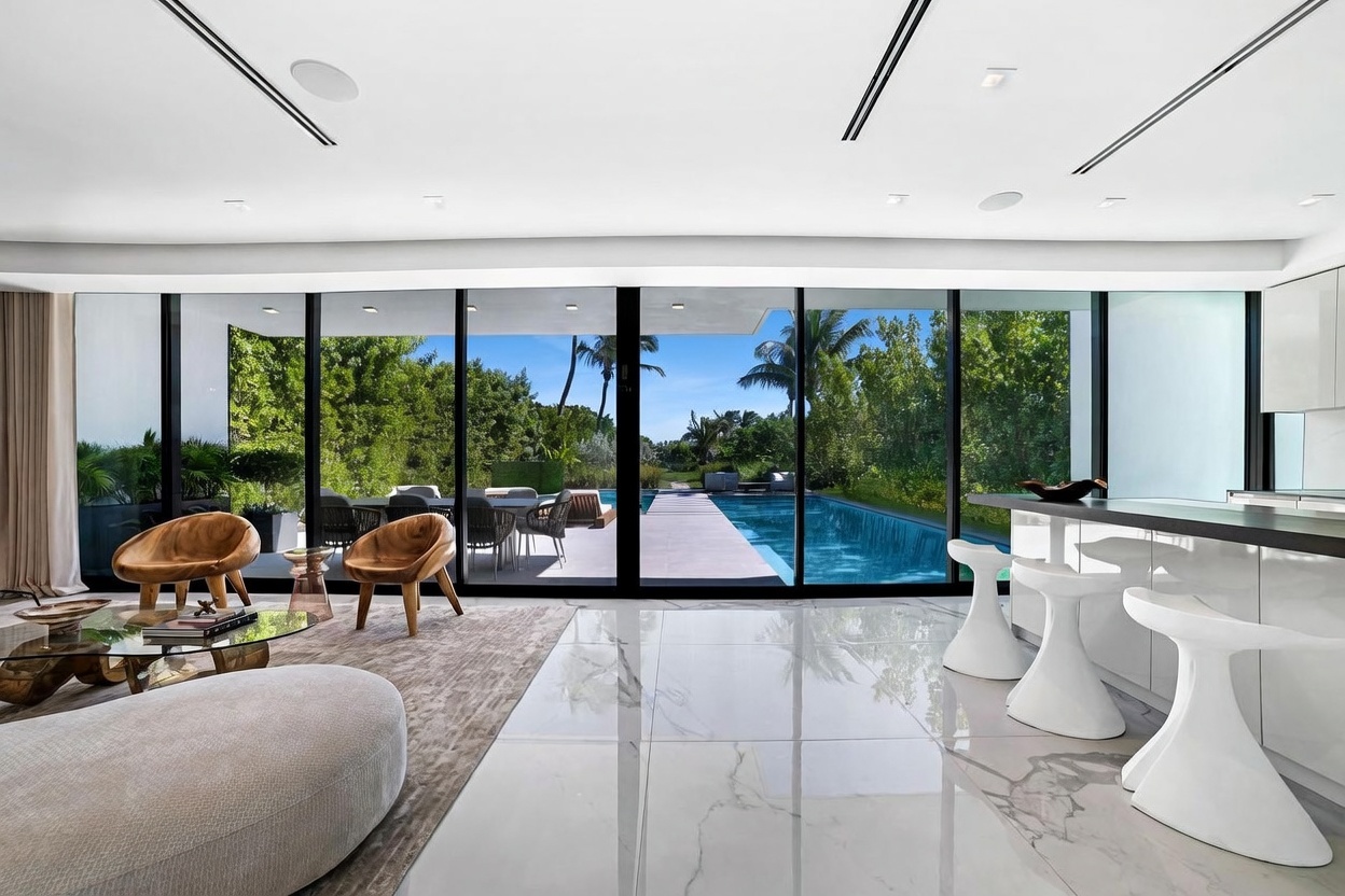 This modern, open-concept living space features polished stone flooring and a seamless transition to the outdoors through expansive floor-to-ceiling glass walls. The room is furnished with sculptural wooden chairs and a low-profile coffee table, creating a sophisticated lounge area that overlooks a private swimming pool and lush greenery. The bright, airy atmosphere is enhanced by recessed lighting and a sleek kitchen island with bar seating visible on the right.