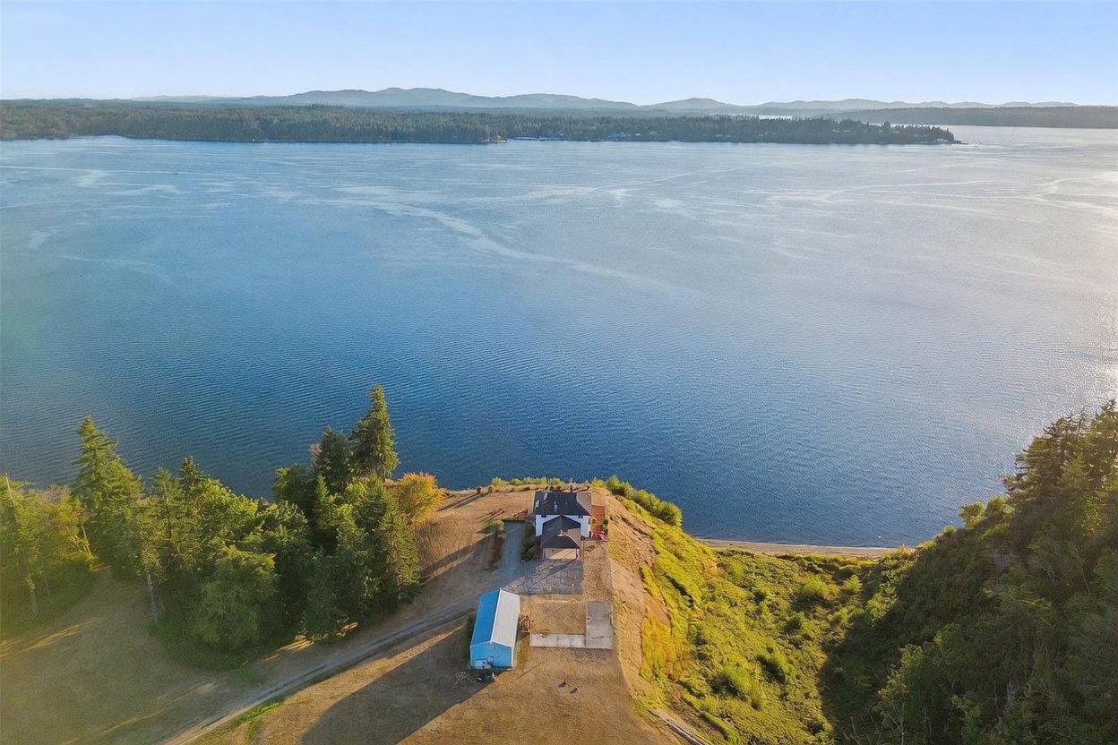 This high-angle aerial view captures a secluded property situated on a prominent bluff overlooking a vast, calm body of water. The site features a main residence, a detached outbuilding, and a gravel driveway, all surrounded by mature trees and open grassy areas that slope down toward the shoreline. The expansive water view and elevated position provide a sense of privacy and tranquility, highlighting the unique coastal landscape.