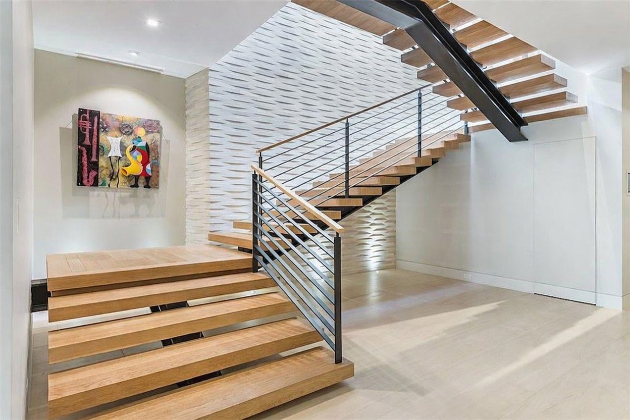 This striking entryway features a modern floating staircase with light wood treads and a sleek black metal railing. A textured, three-dimensional white wall panel serves as a dramatic backdrop, while a vibrant piece of abstract art adds a pop of color to the adjacent neutral wall. The perspective is captured from the base of the stairs, emphasizing the open, airy design and the clean, contemporary aesthetic of the home.