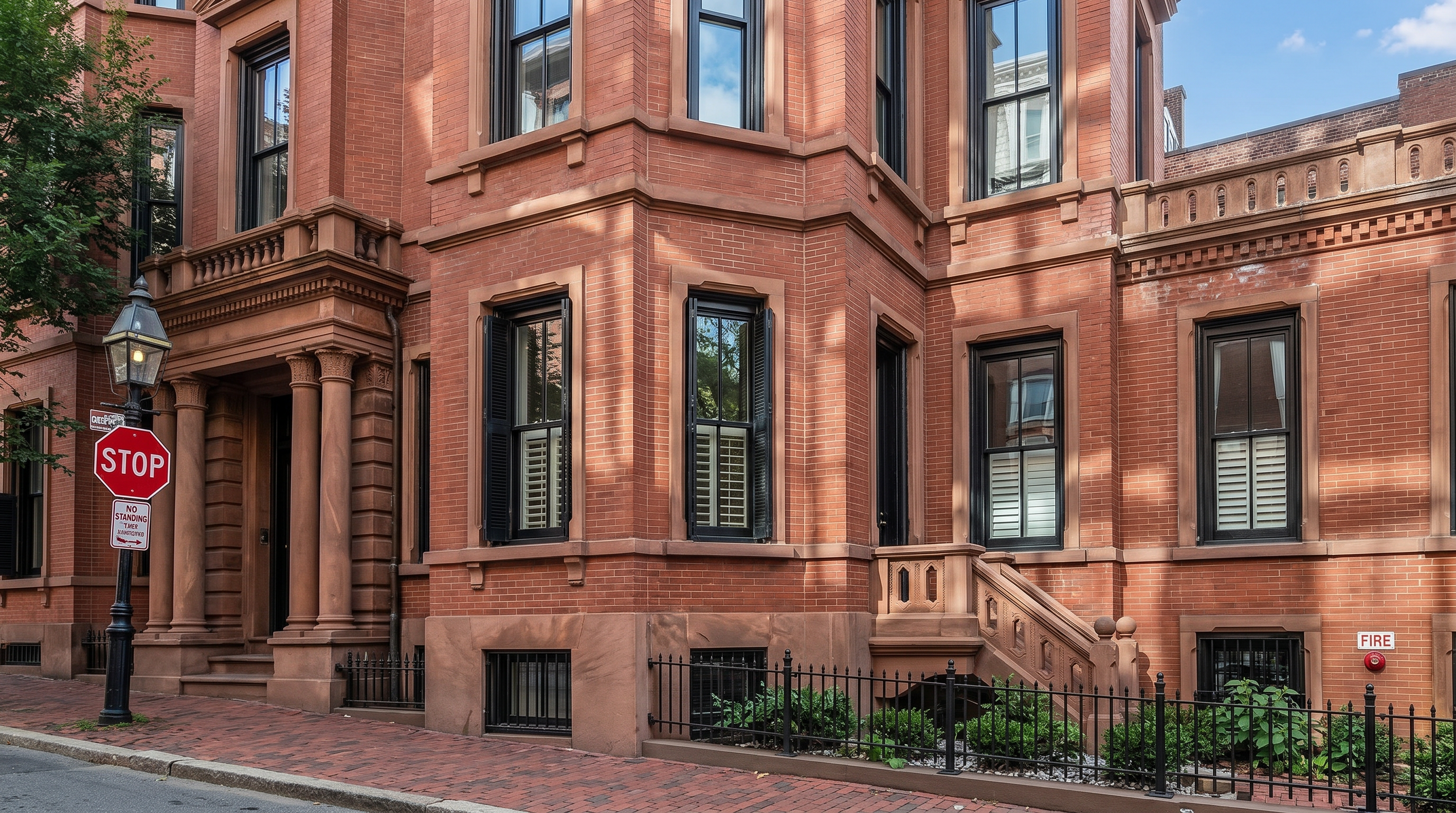 This image captures a stately, multi-story brick townhouse from a low-angle street perspective, emphasizing its classic architectural character. The building features a prominent bay window structure, a mansard-style roofline with dormer windows, and a decorative iron fence lining the sidewalk. The warm red brick facade is complemented by dark-framed windows and traditional masonry detailing, presenting a sophisticated and historic urban aesthetic.
