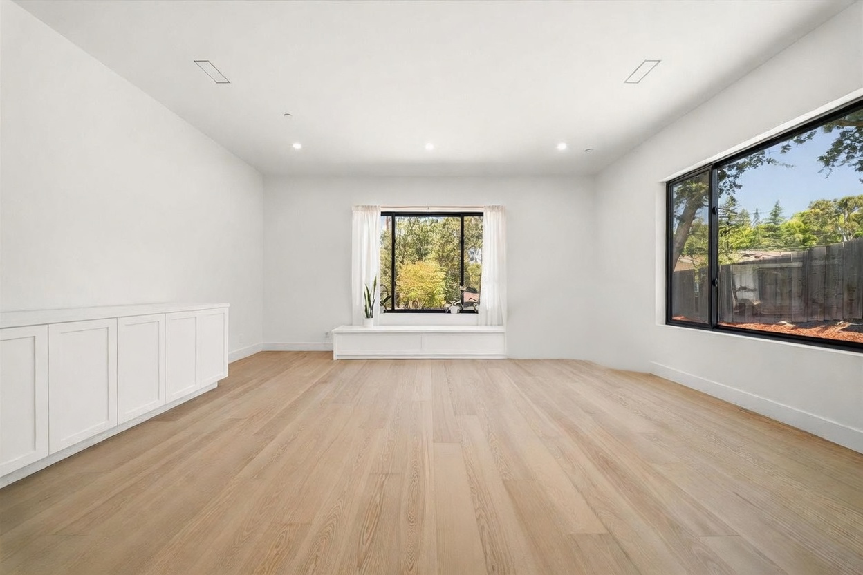 This bright and airy living room features light-toned wood flooring and crisp white walls, creating a clean, minimalist aesthetic. A built-in storage unit lines the left wall, while a low-profile bench sits beneath a central window that looks out onto lush greenery. Large windows on the right wall provide ample natural light and a view of the backyard, contributing to an open and inviting atmosphere.