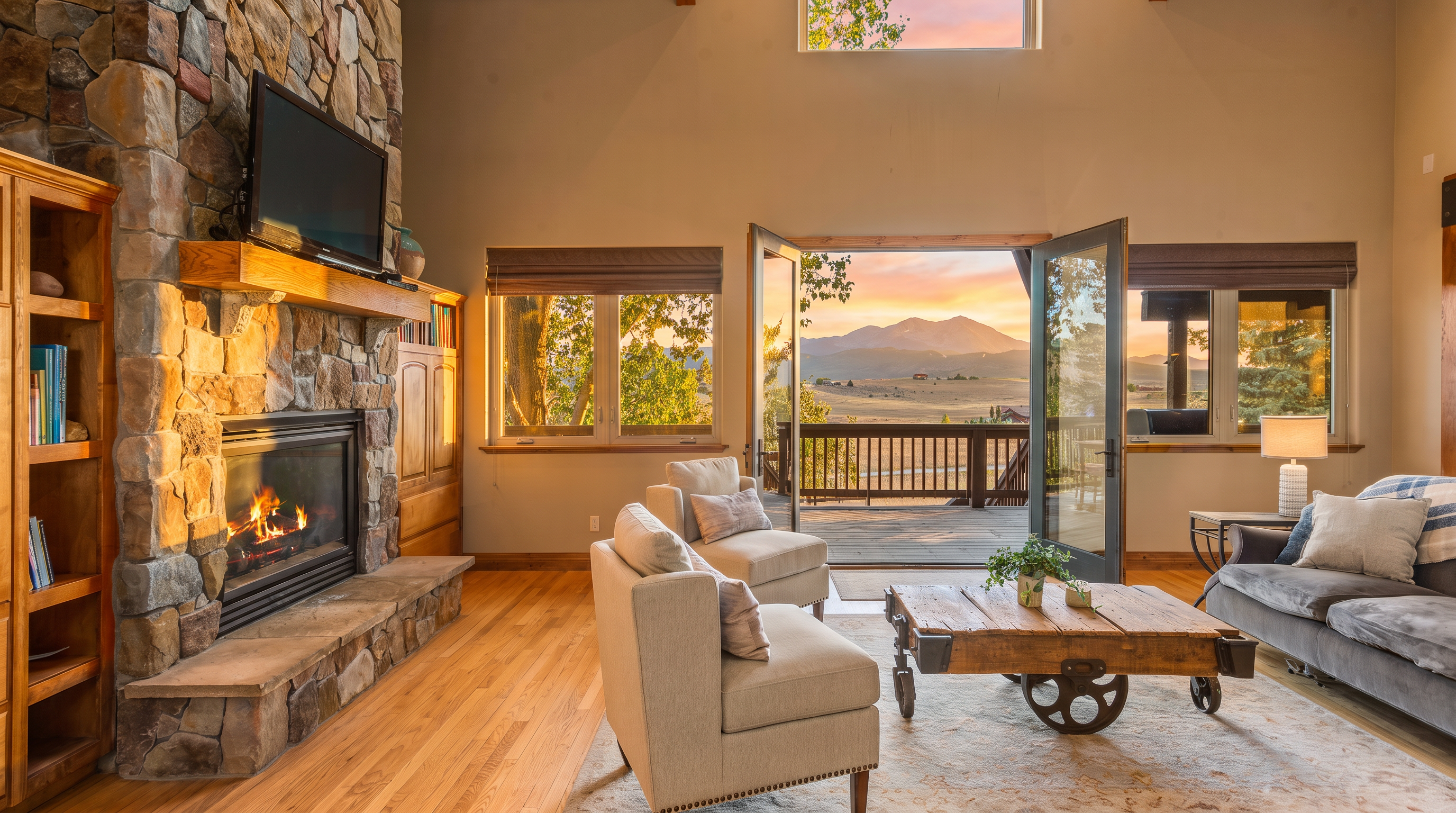 This inviting living room features a floor-to-ceiling stone fireplace as its centerpiece, complemented by a cozy seating area and warm hardwood flooring. Large glass double doors open to a spacious deck, offering breathtaking, unobstructed views of the surrounding mountainous landscape. The room is enhanced by vaulted ceilings with exposed wood beams and thoughtful rustic decor, creating a perfect balance of comfort and natural beauty.