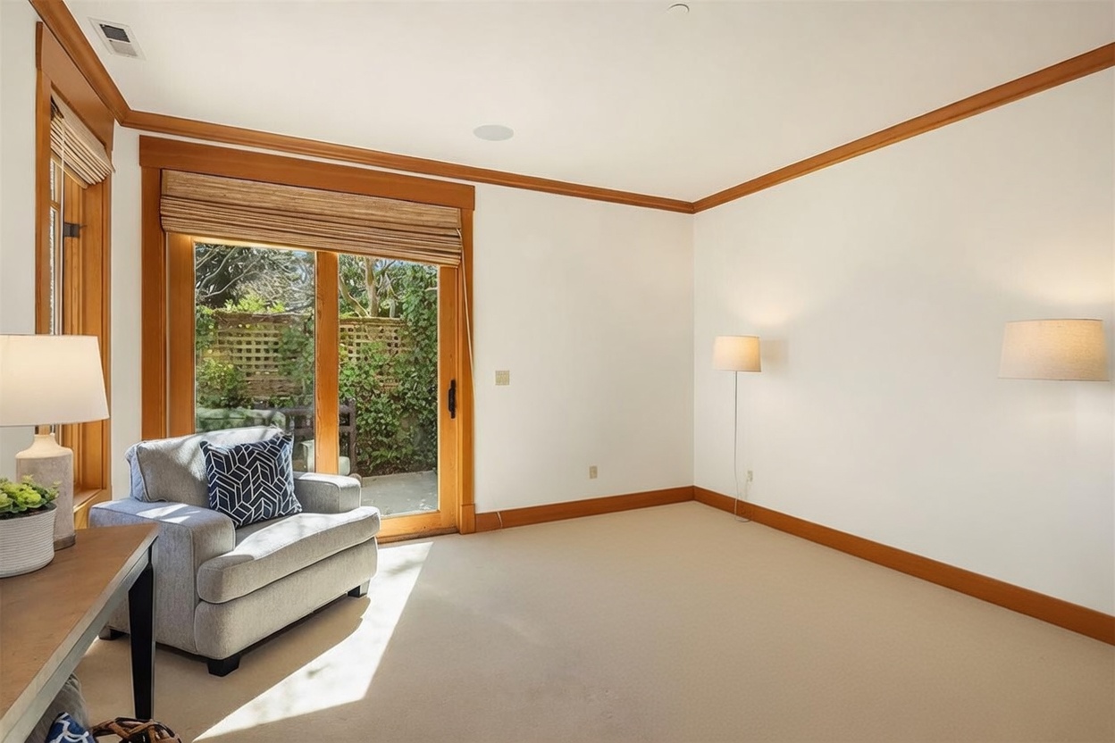 This bright and inviting bedroom features a neutral color palette, a comfortable armchair, and a large sliding glass door that provides direct access to a private outdoor patio. The room is furnished with a queen-sized bed with a wooden headboard, flanked by two matching nightstands and lamps, creating a cozy and serene atmosphere. The perspective is from the corner of the room, capturing the seamless transition between the indoor living space and the lush greenery visible outside.