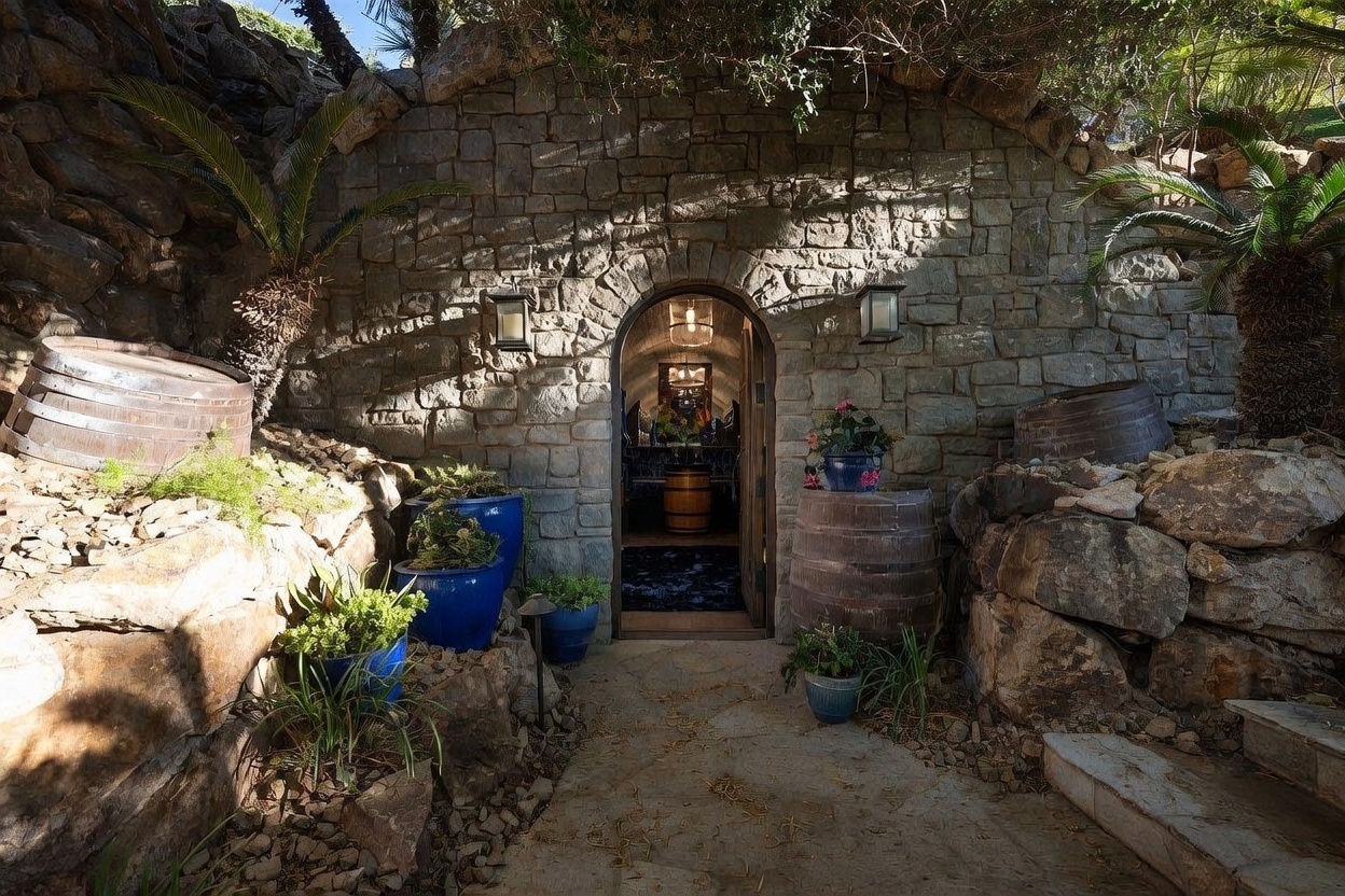 This image captures the rustic, arched stone entryway to a wine cellar, built directly into a hillside. The stone-clad facade features a central arched doorway flanked by decorative wooden barrels and potted plants, creating a Mediterranean-inspired aesthetic. Through the open door, a glimpse of the interior reveals a vaulted, tunnel-like space with a chandelier and additional wine storage, evoking the atmosphere of a traditional European cave cellar.