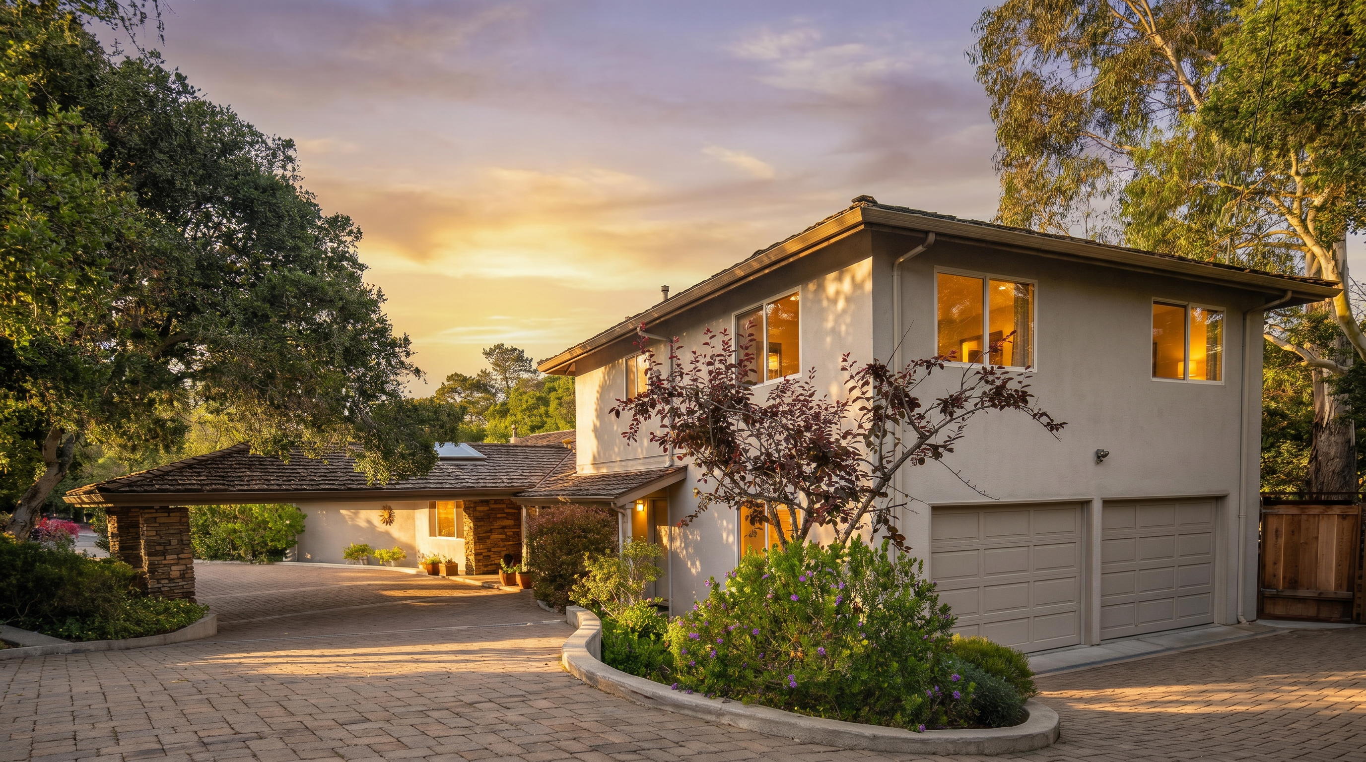 This exterior view captures a two-story home with a light-colored stucco facade and a multi-car garage. A wide, paved driveway leads to a covered carport area supported by stone pillars, while a curved garden bed with vibrant purple flowers adds a touch of color to the foreground. The scene is framed by mature trees under a clear blue sky, presenting a welcoming and well-maintained suburban property.