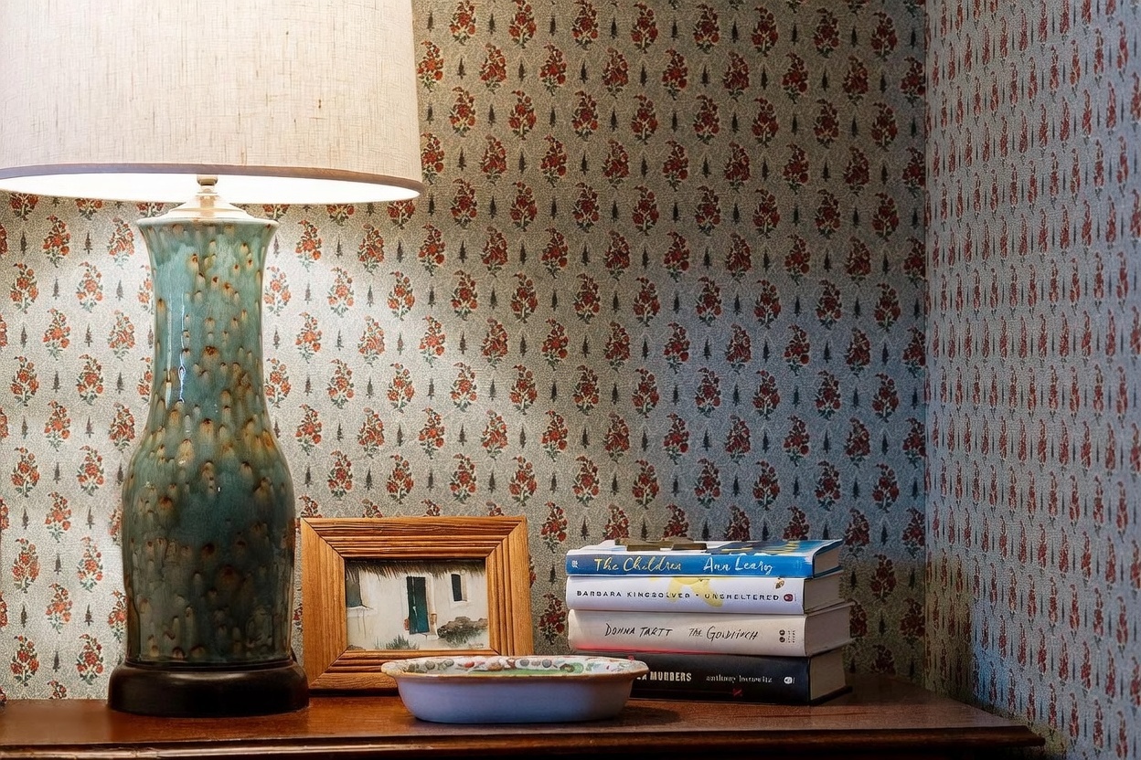 This close-up shot captures a cozy, curated vignette on a wooden surface, featuring a textured, blue-green ceramic table lamp with a classic drum shade. Beside the lamp sits a small framed landscape painting, a shallow decorative bowl, and a stack of four hardcover books, including titles by Ann Leary, Barbara Kingsolver, Donna Tartt, and Anthony Horowitz. The scene is set against a backdrop of traditional, floral-patterned wallpaper, creating a warm and inviting atmosphere.