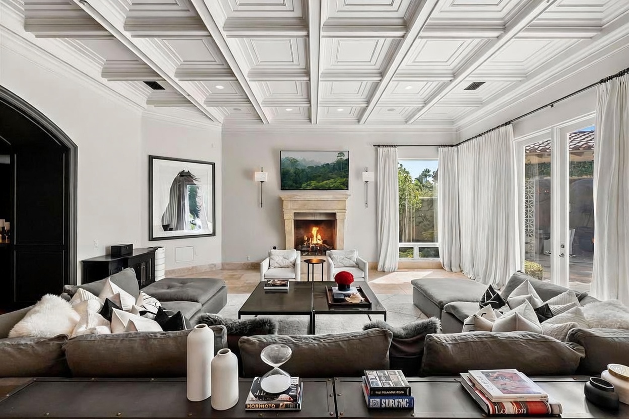 This is a luxurious living room featuring a coffered ceiling, a stone fireplace with a mounted television, and comfortable, oversized sofas. The room is decorated in neutral tones with pops of black and white, creating a sophisticated and inviting atmosphere. Large windows with sheer curtains provide ample natural light and a view to the outside.