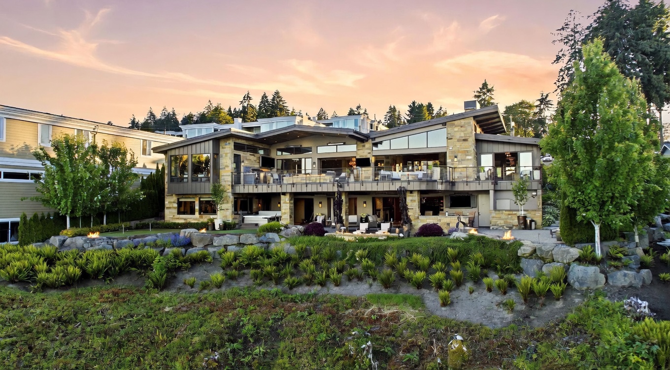 This expansive rear view of a contemporary multi-level home showcases a sophisticated architectural design with stone and wood siding. The property features a large elevated deck with outdoor seating and a lower-level patio area complete with a hot tub and fire pit, all set against a beautifully landscaped terraced yard. The warm sunset lighting highlights the home's modern aesthetic and seamless transition between indoor and outdoor living spaces.