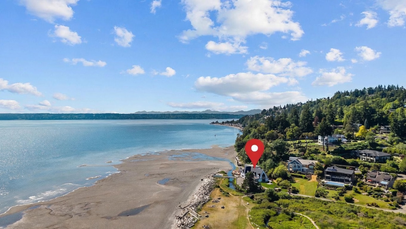 This high-angle aerial shot captures a waterfront property nestled along a scenic coastline, featuring a prominent house marked by a red location pin. The landscape includes a sandy beach with driftwood, lush green trees, and a clear view of the expansive bay under a bright, partly cloudy sky. The perspective emphasizes the home's secluded, prime location with direct access to the shoreline.