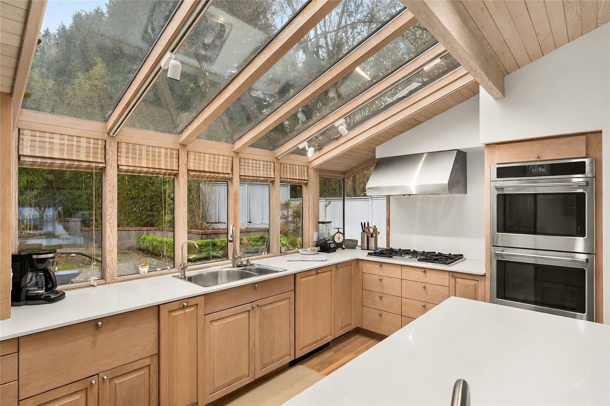 This bright, contemporary kitchen features light wood cabinetry, white countertops, and a prominent stainless steel double wall oven. A unique sloped glass ceiling and large windows provide an abundance of natural light and a clear view of the outdoor greenery. The perspective is from the kitchen island, looking toward the sink and cooking area, creating an airy and inviting atmosphere.