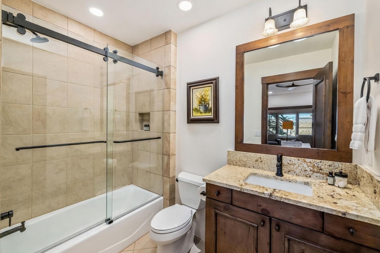 This bathroom features a vanity with a stone countertop, a dark wood cabinet, and a framed mirror reflecting a view into an adjacent room. To the left, a glass-enclosed shower and tub combination is finished with neutral-toned wall tiles and includes a built-in recessed niche. The space is illuminated by a dual-light fixture above the mirror and recessed ceiling lights, creating a warm and functional atmosphere.