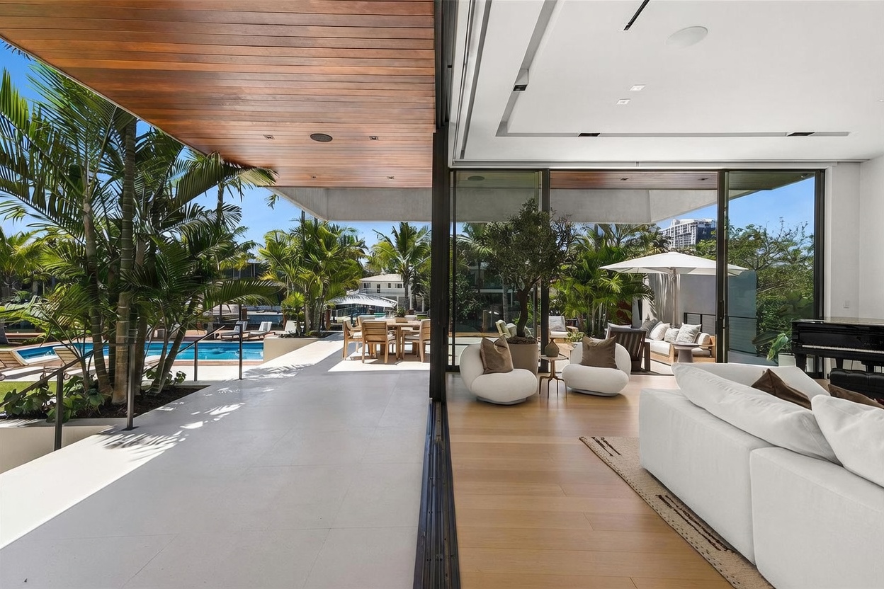 This image captures a seamless transition between a bright, modern living room and an expansive outdoor patio, facilitated by large sliding glass doors. The interior features light wood flooring and a plush white sofa, while the covered patio area offers a dining space overlooking a swimming pool and lush tropical landscaping. The perspective emphasizes an open-concept design that blurs the lines between indoor and outdoor living, creating a serene and luxurious atmosphere.