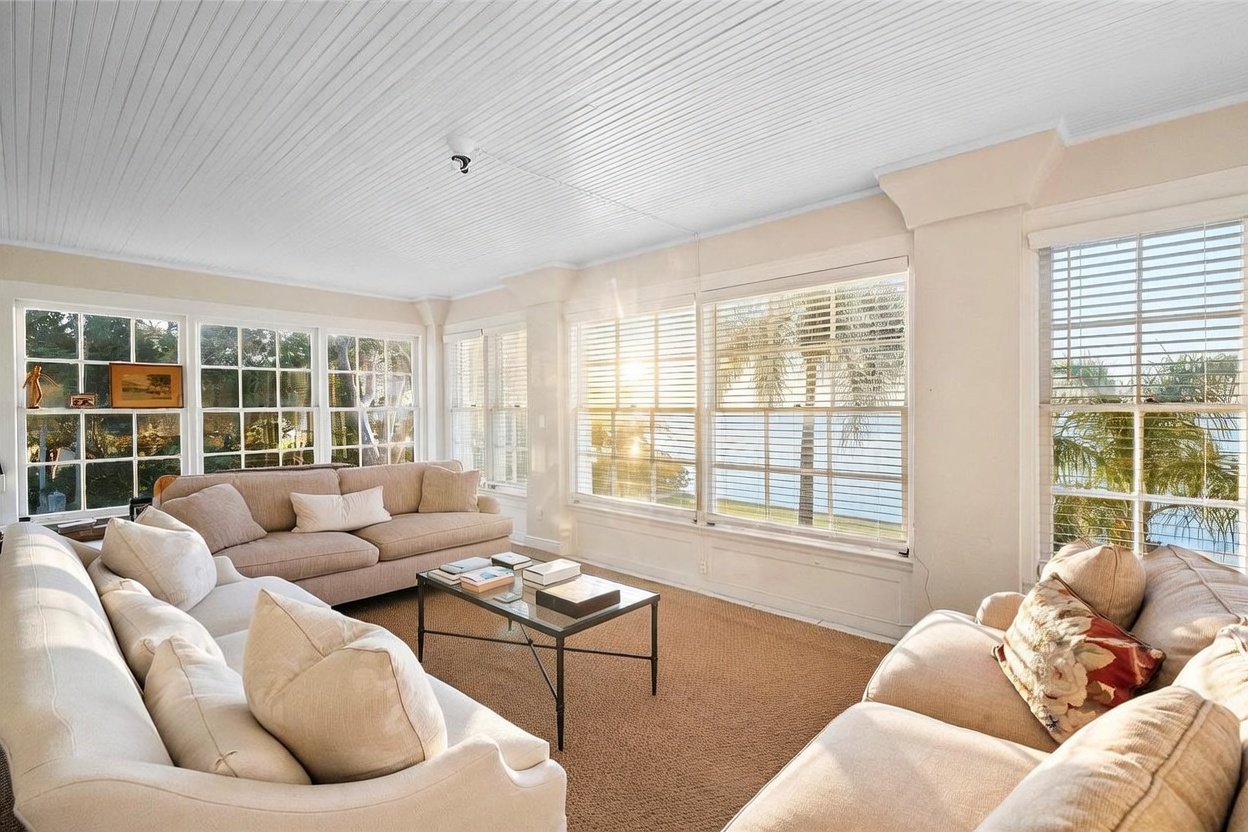 This bright and airy sunroom features a neutral color palette with two plush sofas arranged around a central glass-topped coffee table. The space is defined by expansive walls of windows that offer panoramic views of the water and surrounding greenery, allowing for abundant natural light. The room is finished with a textured area rug and a white paneled ceiling, creating a relaxed and inviting atmosphere.