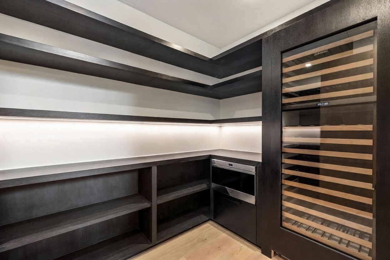 This sophisticated walk-in pantry features dark, custom-built cabinetry and open shelving that wraps around the corner. The space is equipped with a built-in microwave and a tall, glass-fronted wine refrigerator, all illuminated by integrated under-shelf lighting. The light wood flooring provides a warm contrast to the dark cabinetry, creating a modern and organized storage area.