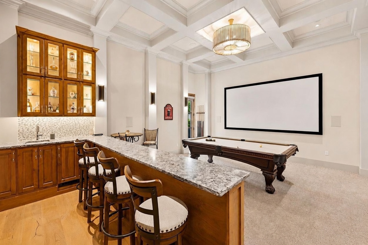 This spacious entertainment area features a built-in wet bar with glass-front cabinetry and a stone-topped counter, complemented by three bar stools. A pool table sits in the center of the room, positioned in front of a large projection screen mounted on the wall. The room is finished with a coffered ceiling, recessed lighting, and a decorative chandelier, creating a sophisticated atmosphere for recreation.