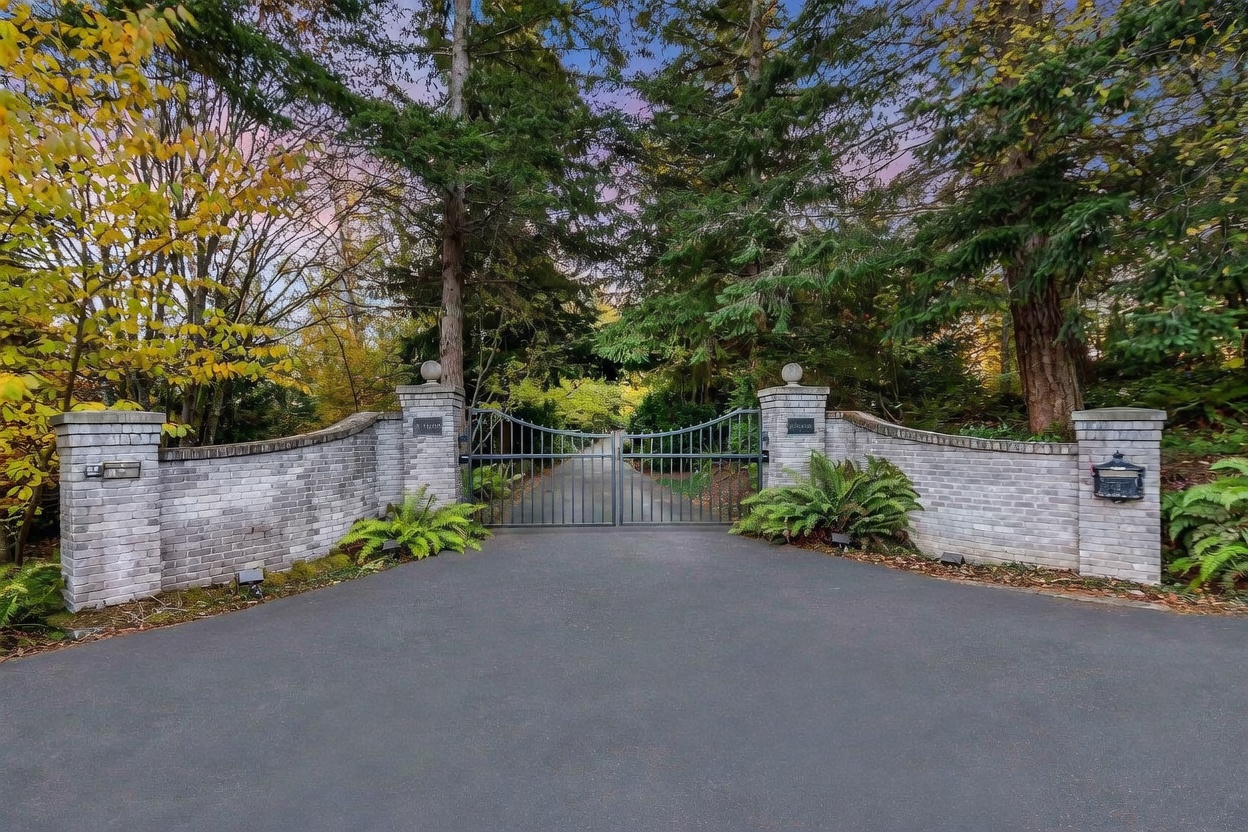 This image captures the grand gated entrance to a private property, featuring a symmetrical design with light-colored brick pillars and curved walls. A dark metal gate stands closed between the pillars, flanked by lush green ferns and mature trees that create a sense of seclusion and privacy. The perspective is a straight-on, eye-level shot that emphasizes the stately and secure nature of the estate's approach.