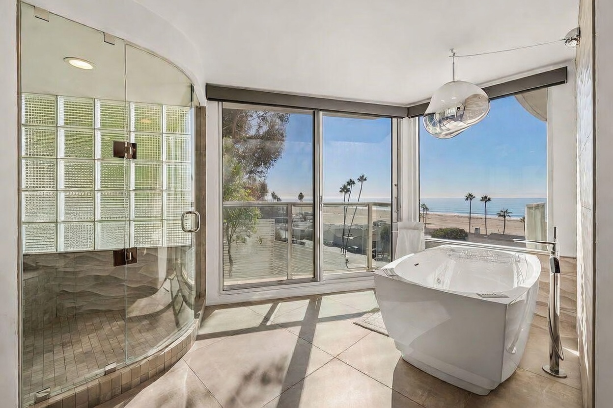 This luxurious primary bathroom features a deep soaking tub positioned to take advantage of expansive ocean views through large sliding glass doors. To the left, a walk-in shower is enclosed by glass panels and accented with a decorative glass-block wall. The space is finished with neutral floor tiles and a modern, spherical pendant light, creating a serene and high-end coastal atmosphere.
