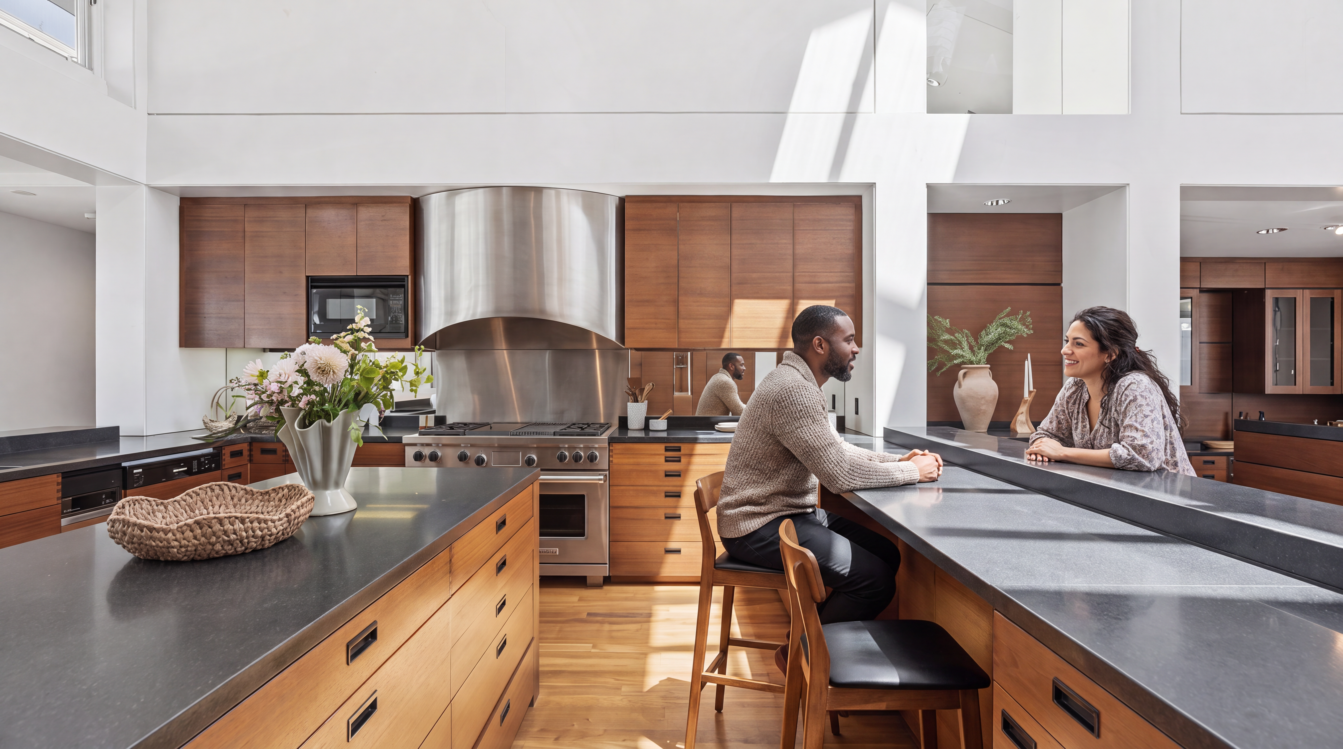 This sleek, modern kitchen features warm wood cabinetry contrasted with dark stone countertops and high-end stainless steel appliances, including a professional-grade range and hood. A large central island with a woven basket and floral arrangement serves as a natural focal point, while the breakfast bar creates an inviting space for social interaction. The bright, open-plan layout is bathed in natural light, giving the room an airy and sophisticated feel.