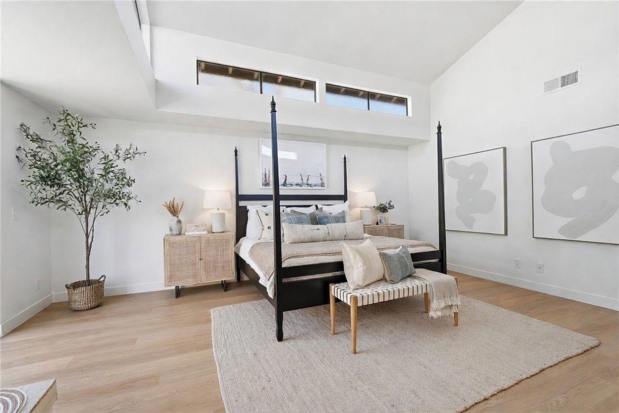 This spacious primary bedroom features high vaulted ceilings with clerestory windows that flood the room with natural light. A black four-poster bed serves as the focal point, complemented by light wood flooring, a textured area rug, and a wooden bench at the foot of the bed. The room is decorated with minimalist art and neutral tones, creating a serene and sophisticated atmosphere.