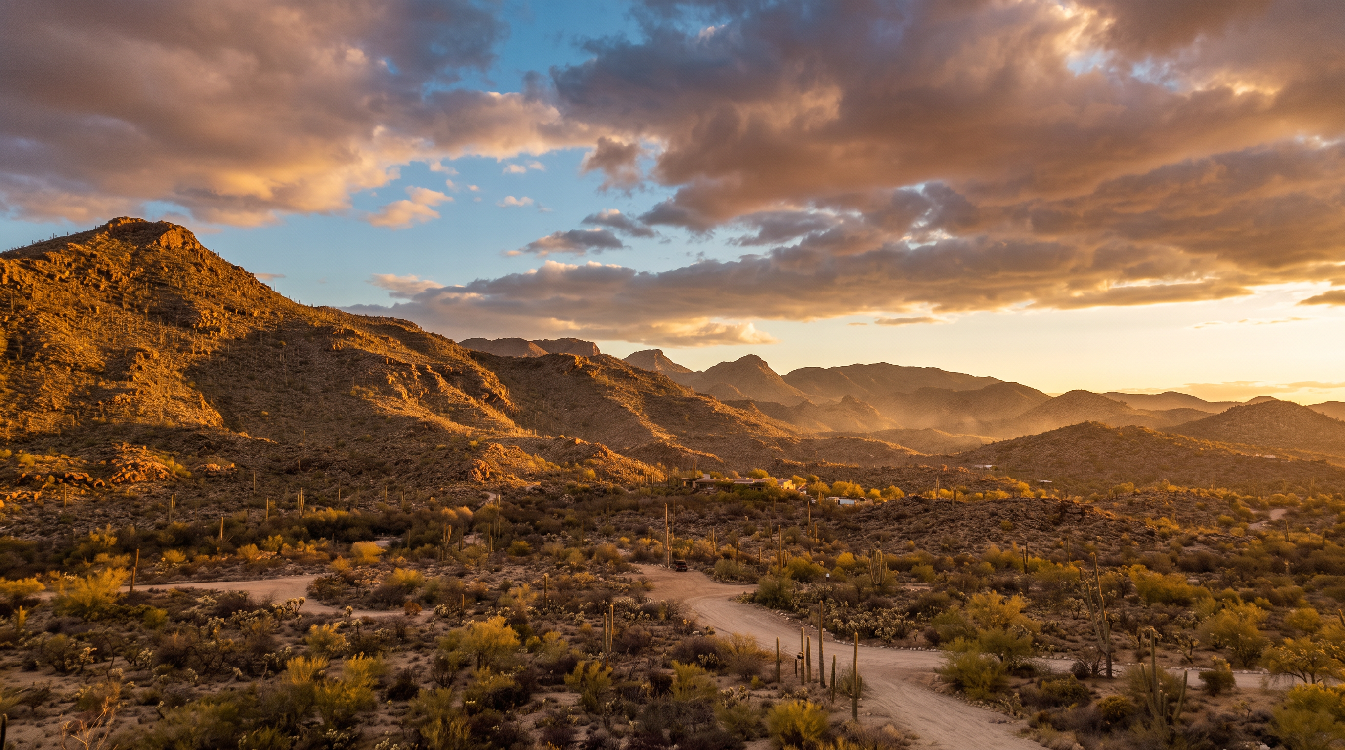 This landscape photograph captures an expansive, rugged desert setting featuring rolling hills and sparse, native vegetation like saguaro cacti. A dirt path winds through the foreground, leading the eye toward the distant mountain range under a dramatic, cloud-filled sky. The image emphasizes the natural beauty, seclusion, and vast outdoor space characteristic of remote desert properties.