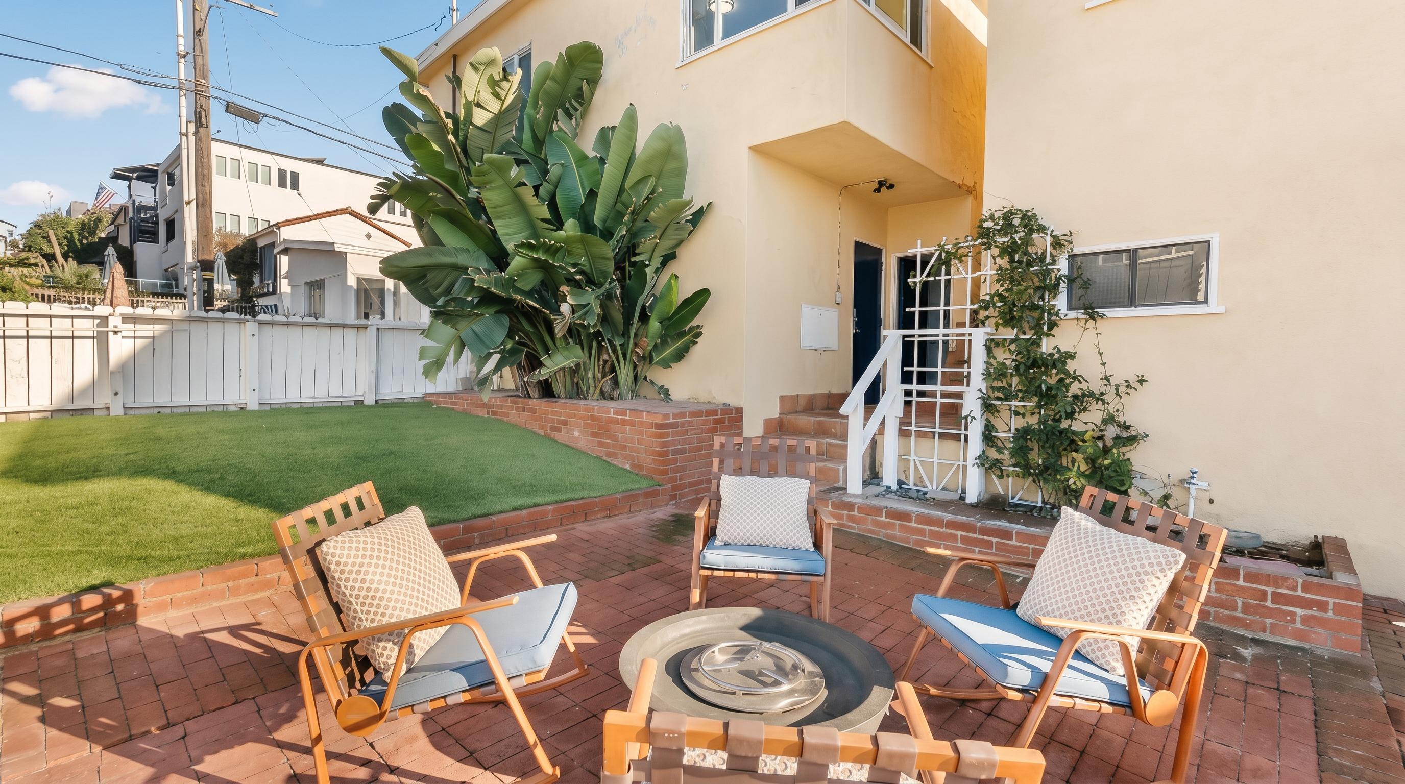 This outdoor living space features a red brick patio arranged with four rocking chairs surrounding a central fire pit. The area is bordered by a raised brick planter containing a large, lush tropical plant and a small patch of artificial turf. A white wooden trellis and a small set of stairs lead toward the building's entrance, creating a cozy and inviting atmosphere for relaxation.